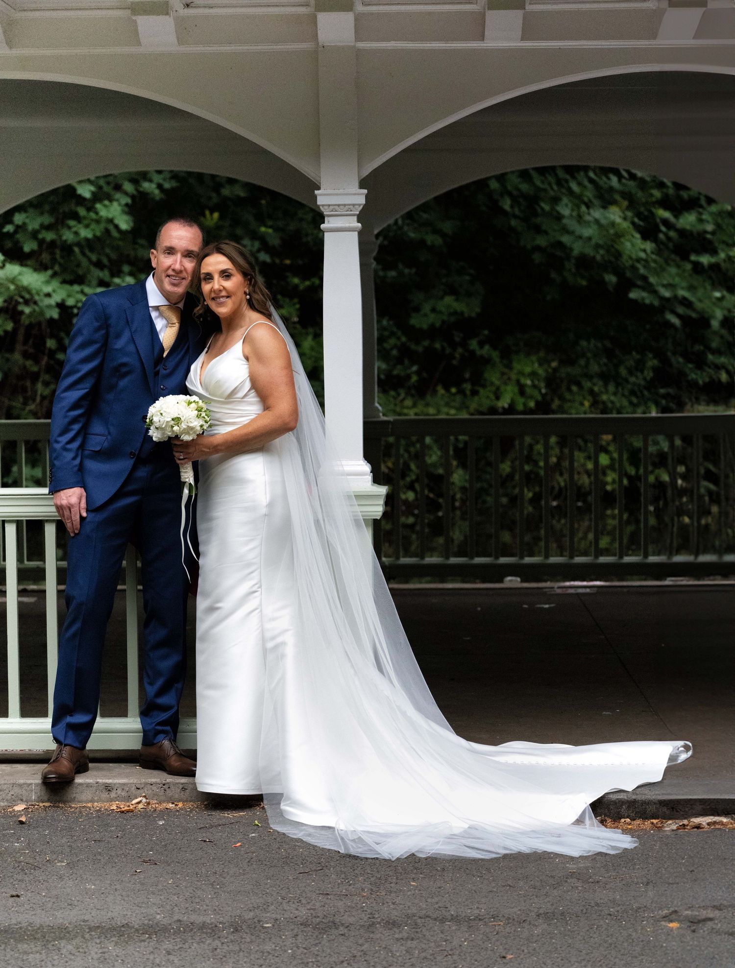 A bride and groom pose elegantly under a white gazebo, she in a white gown with veil, he in a navy suit.