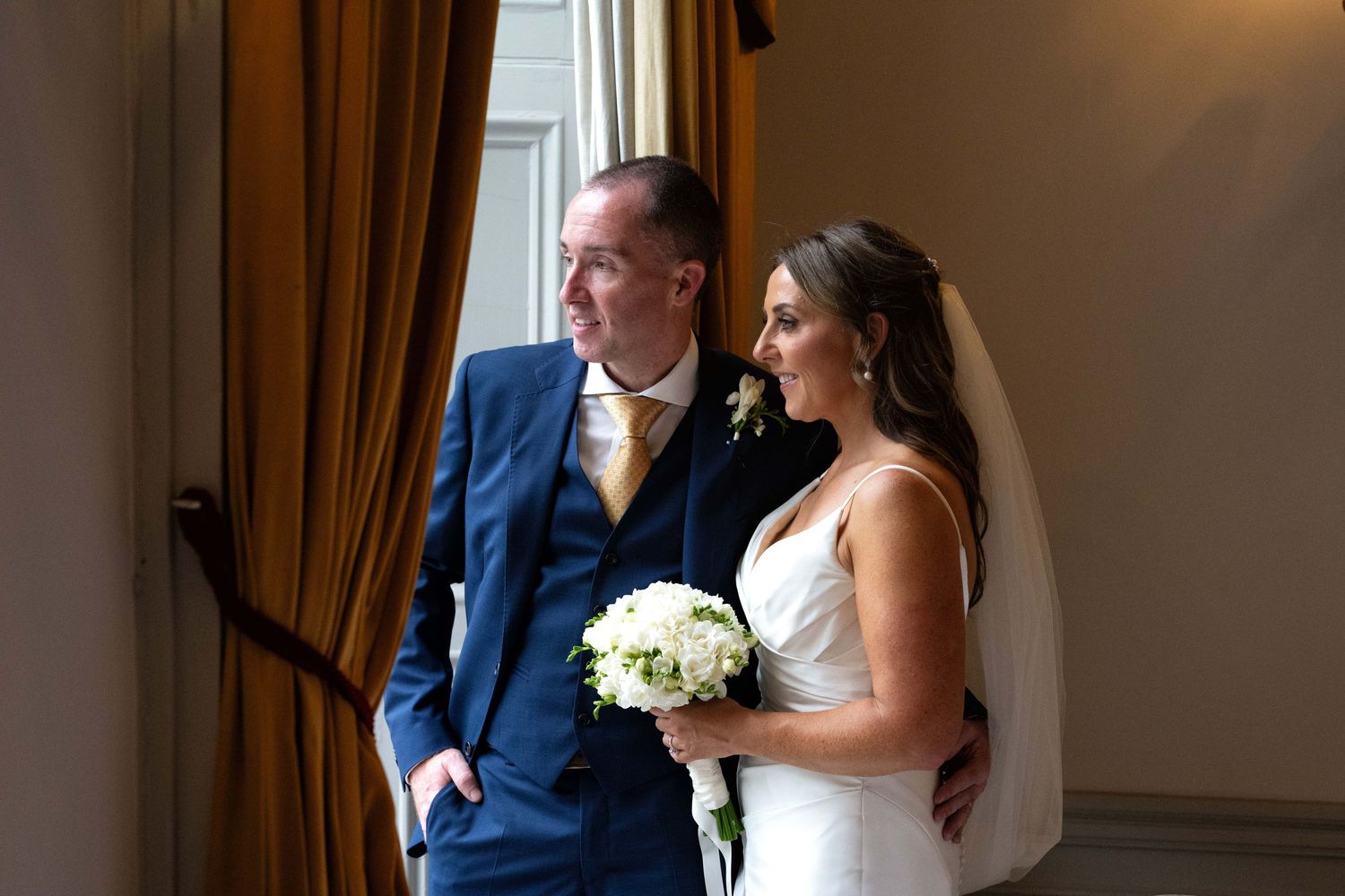 Bride and groom pose elegantly by a window, bride holding white bouquet in a wedding venue.
