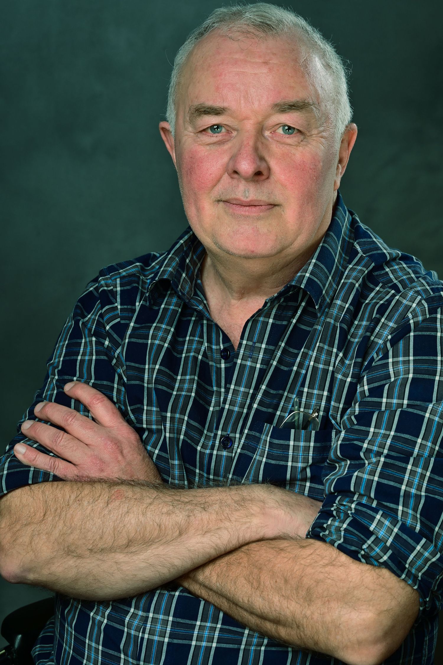 Older man with gray hair, arms crossed, wearing a blue plaid shirt, posing confidently against a dark background.