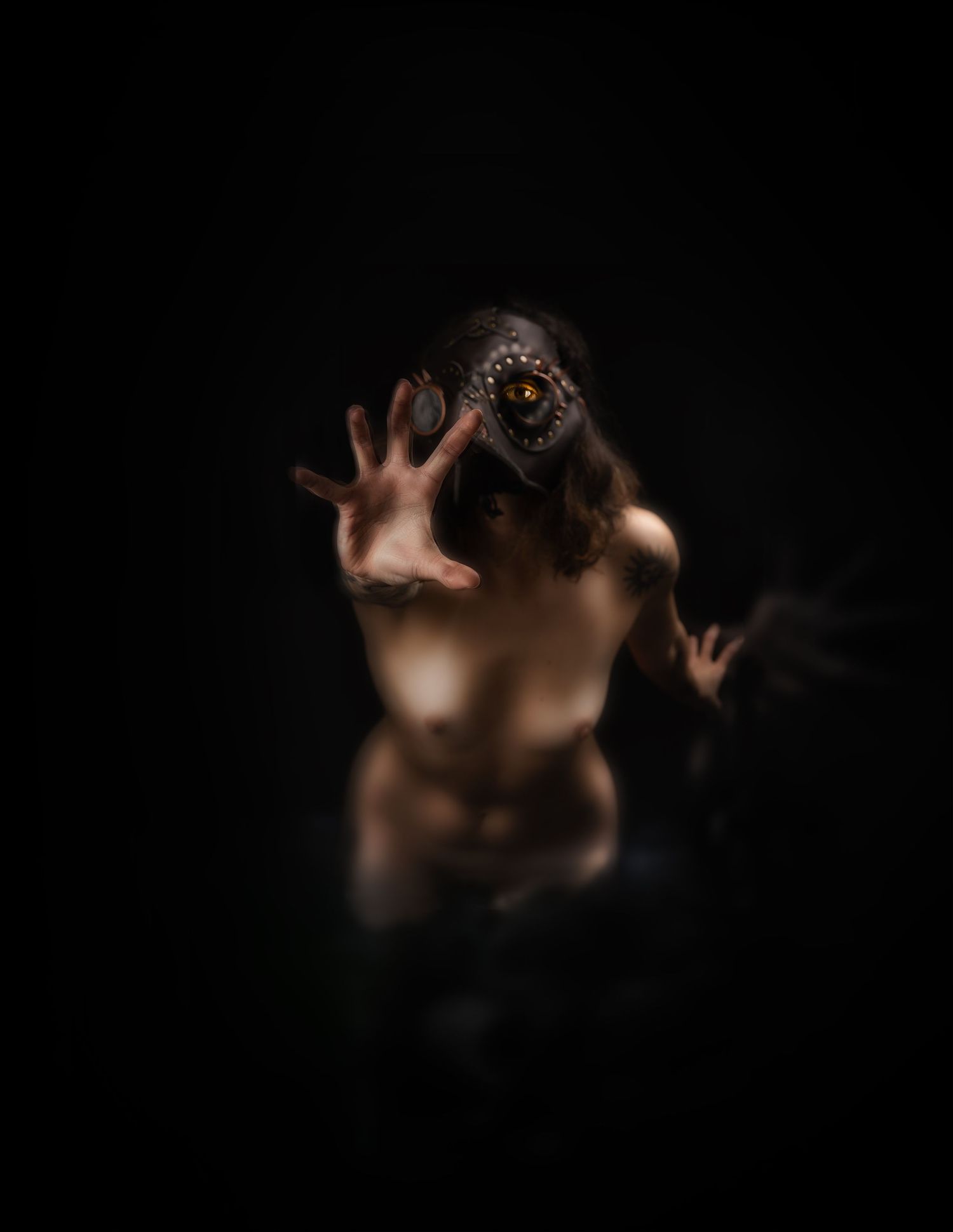 Eerie figure wearing dark bird mask with red eye reaching toward camera in dramatic dark studio photography.