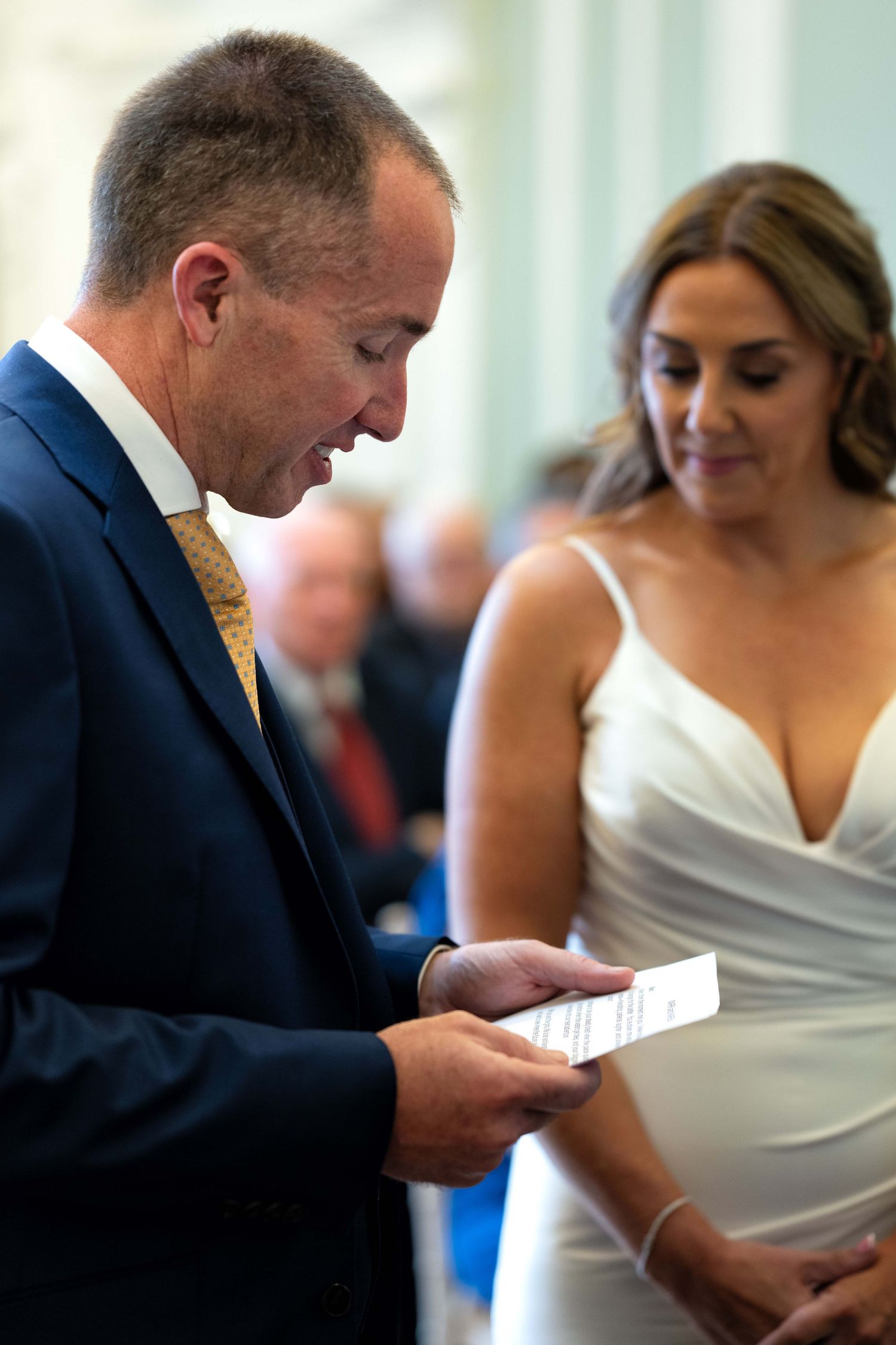 Groom in navy suit reads vows from card while bride in white gown smiles during wedding ceremony.