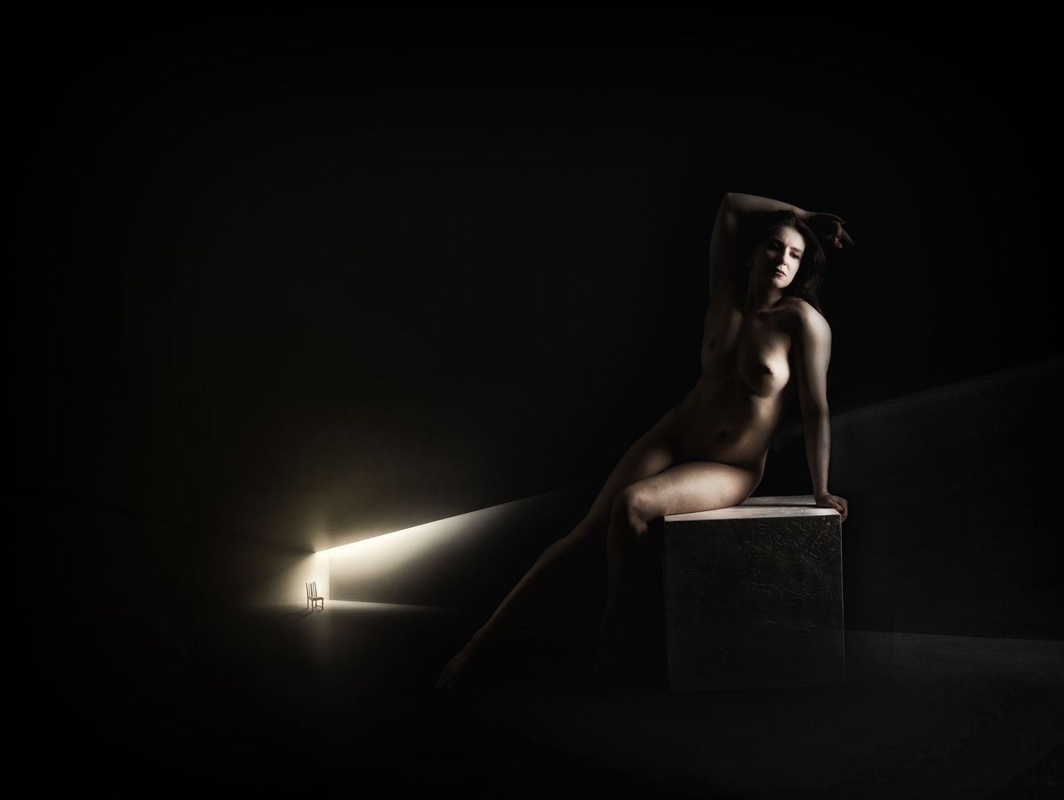 A nude figure sits on a dark box illuminated by dramatic side lighting against a black background.