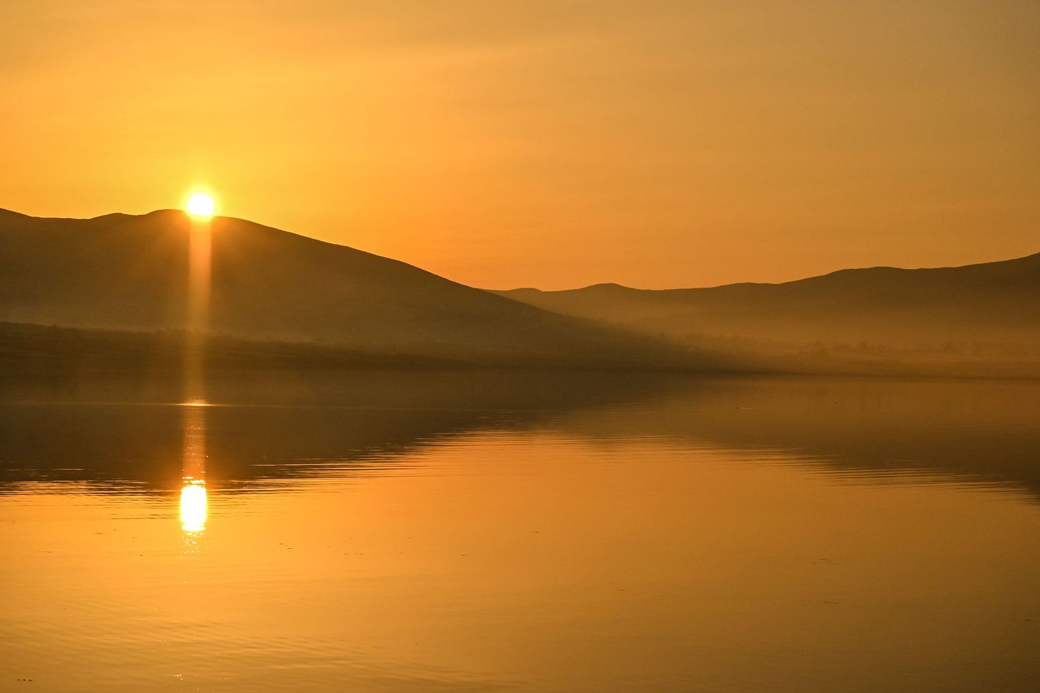 Golden sunrise reflecting on calm lake water with misty mountains silhouetted in the background.