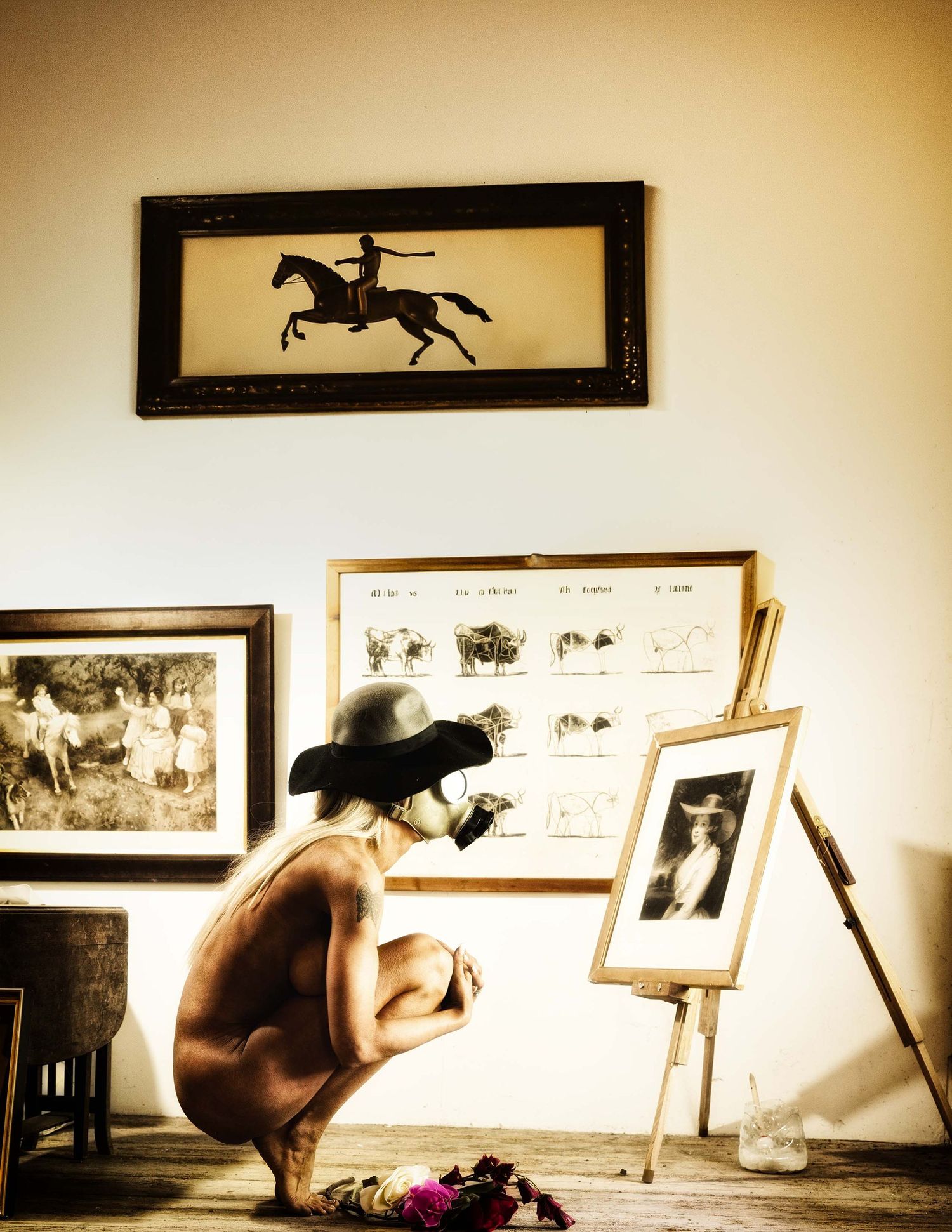 Person crouching before an easel with artwork, surrounded by framed equestrian and vintage photographs on a warm-toned wall.