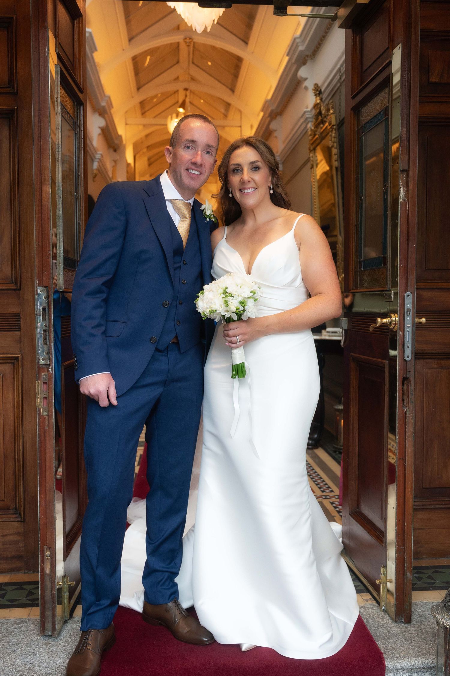 A smiling bride and groom pose in a grand doorway, she in a white gown with bouquet, he in a navy blue suit.