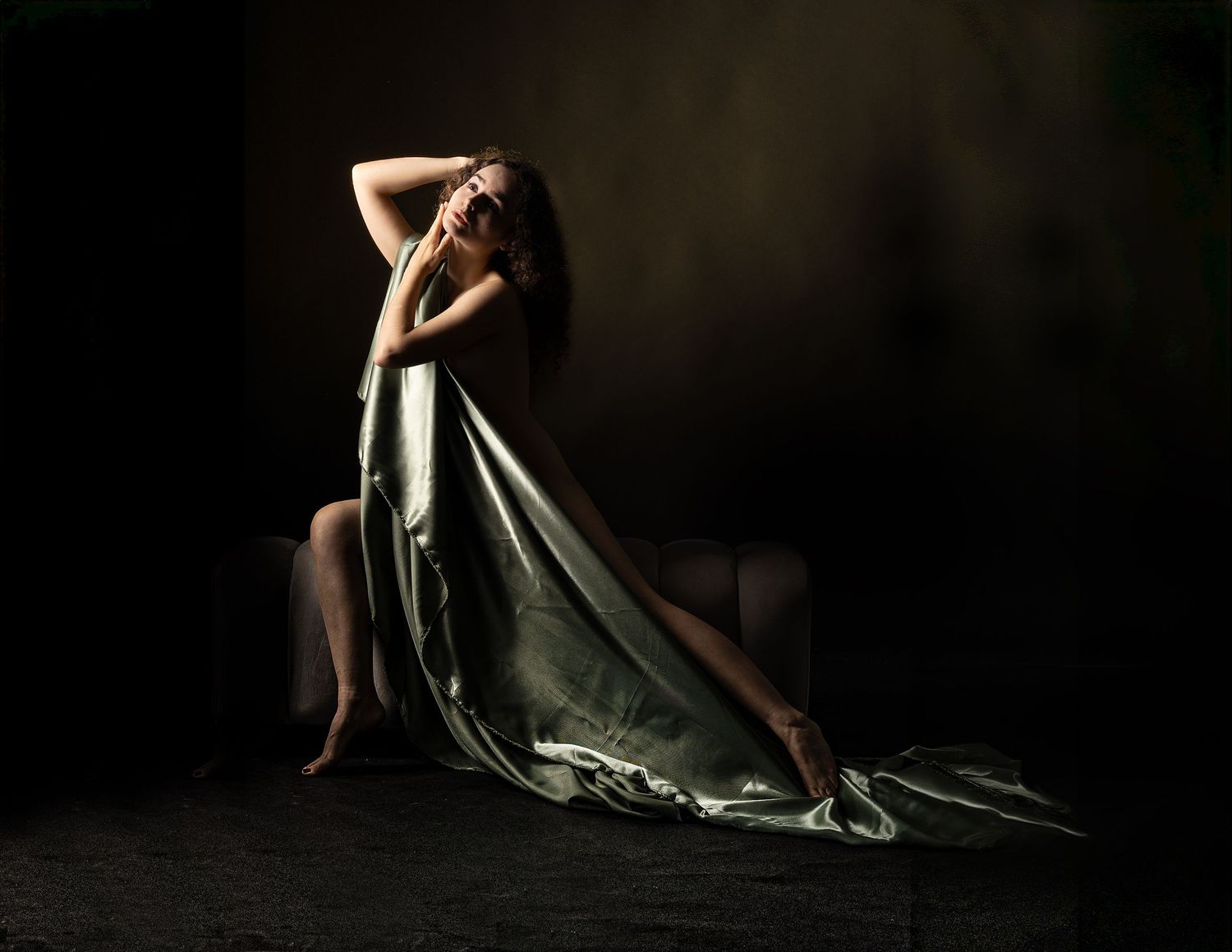 A woman draped in flowing green satin fabric poses gracefully in a dramatic dark studio setting.