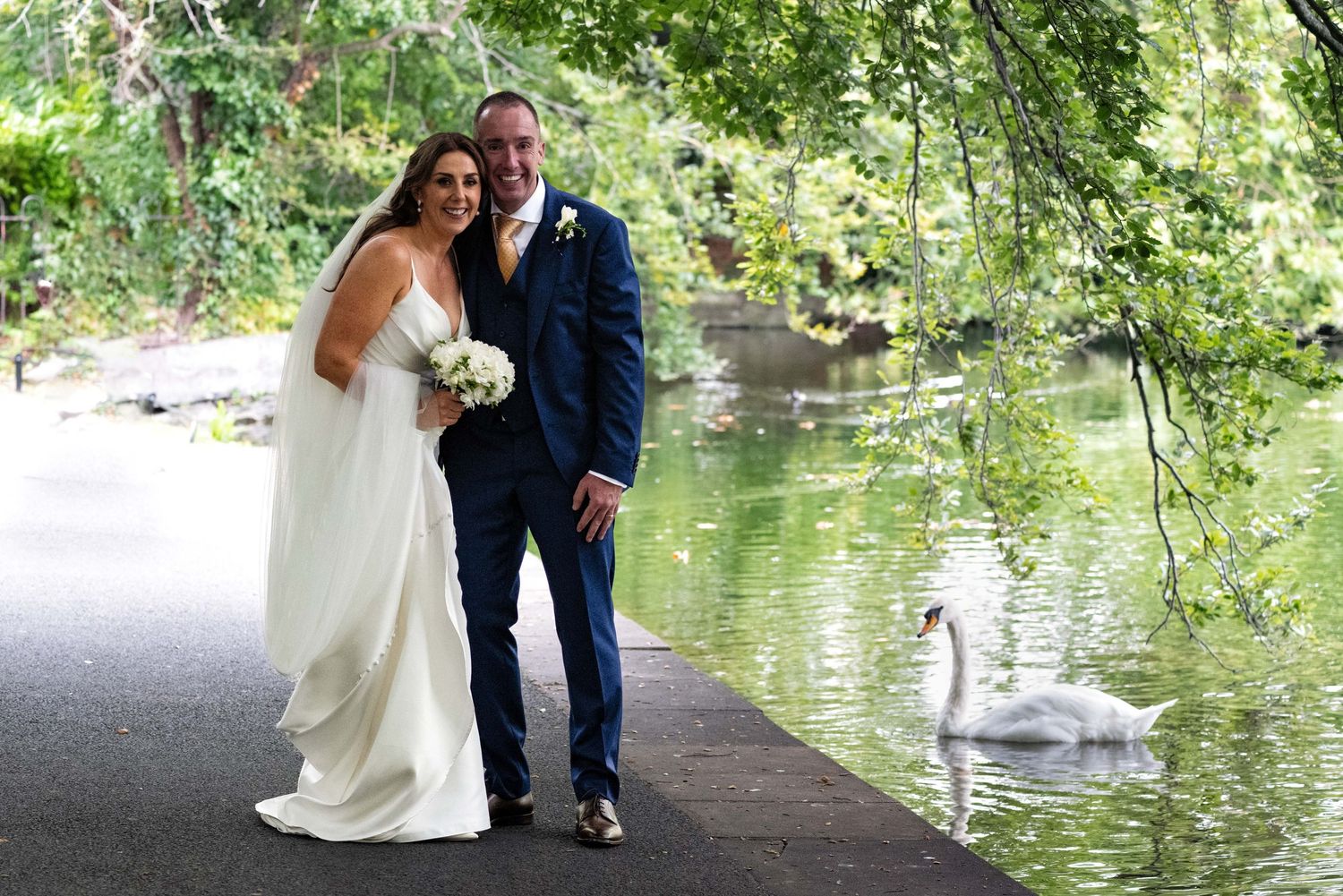 Newlywed couple posing by a tranquil pond with a swan, bride in white gown holding bouquet, groom in navy suit.