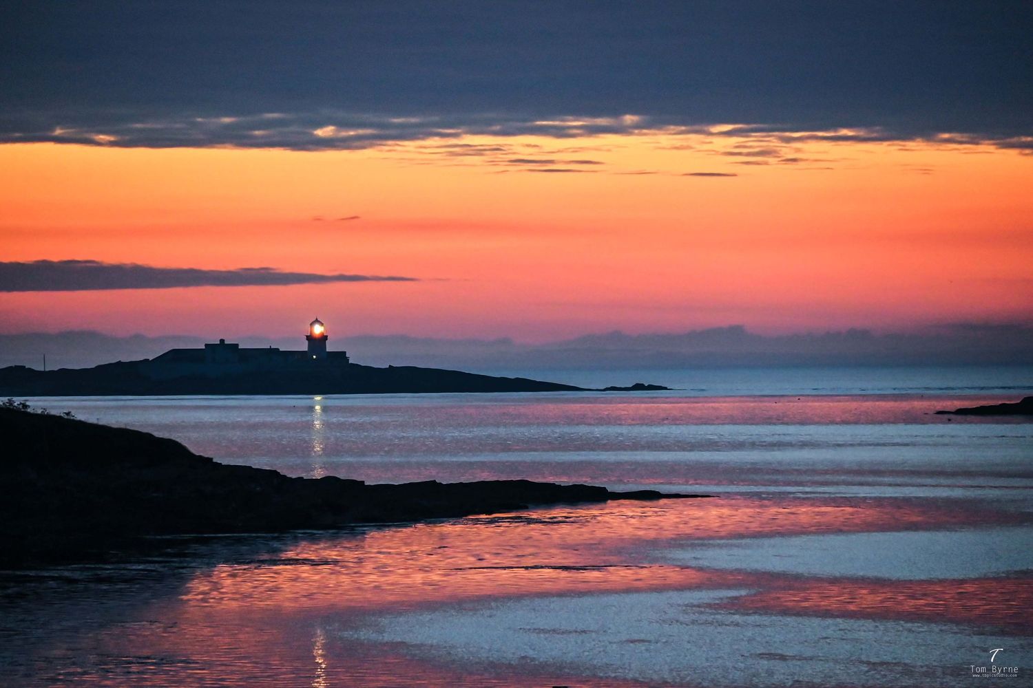 Stunning sunset over calm ocean waters with a glowing lighthouse silhouette on a rocky island under vibrant orange skies.