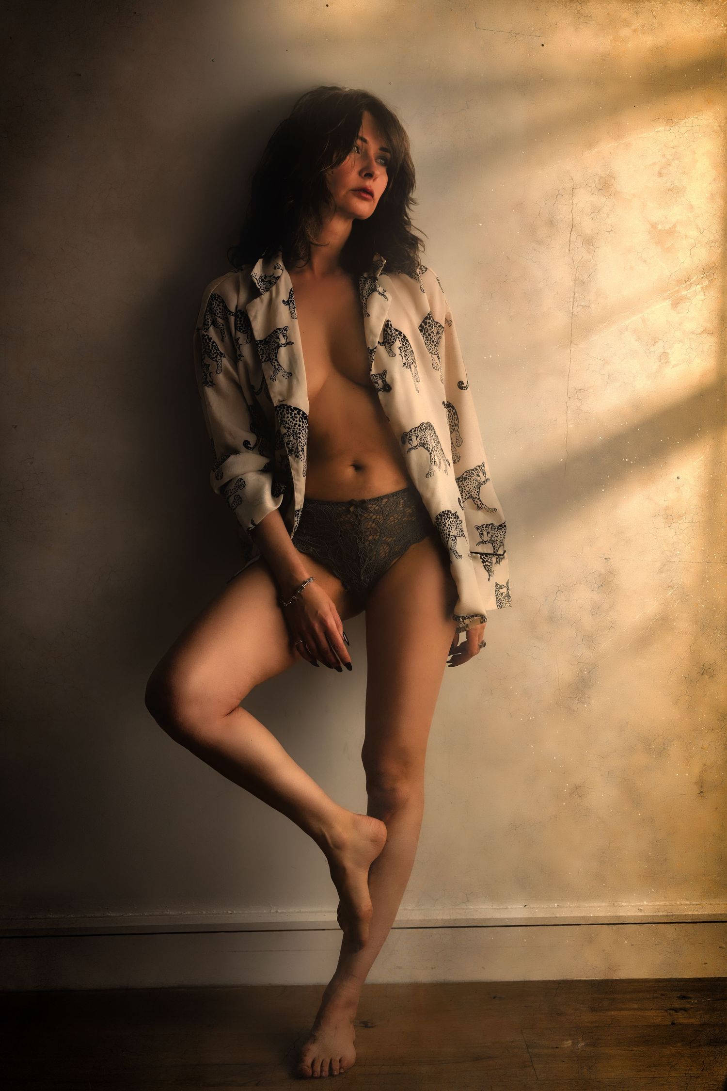 Woman in open printed shirt and lace underwear posing against textured wall in warm dramatic lighting.