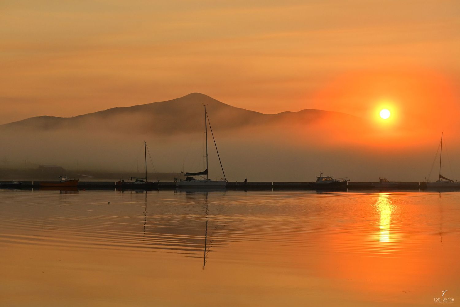 Sailboats silhouetted against a vibrant orange sunset over calm water with misty mountains in the background.