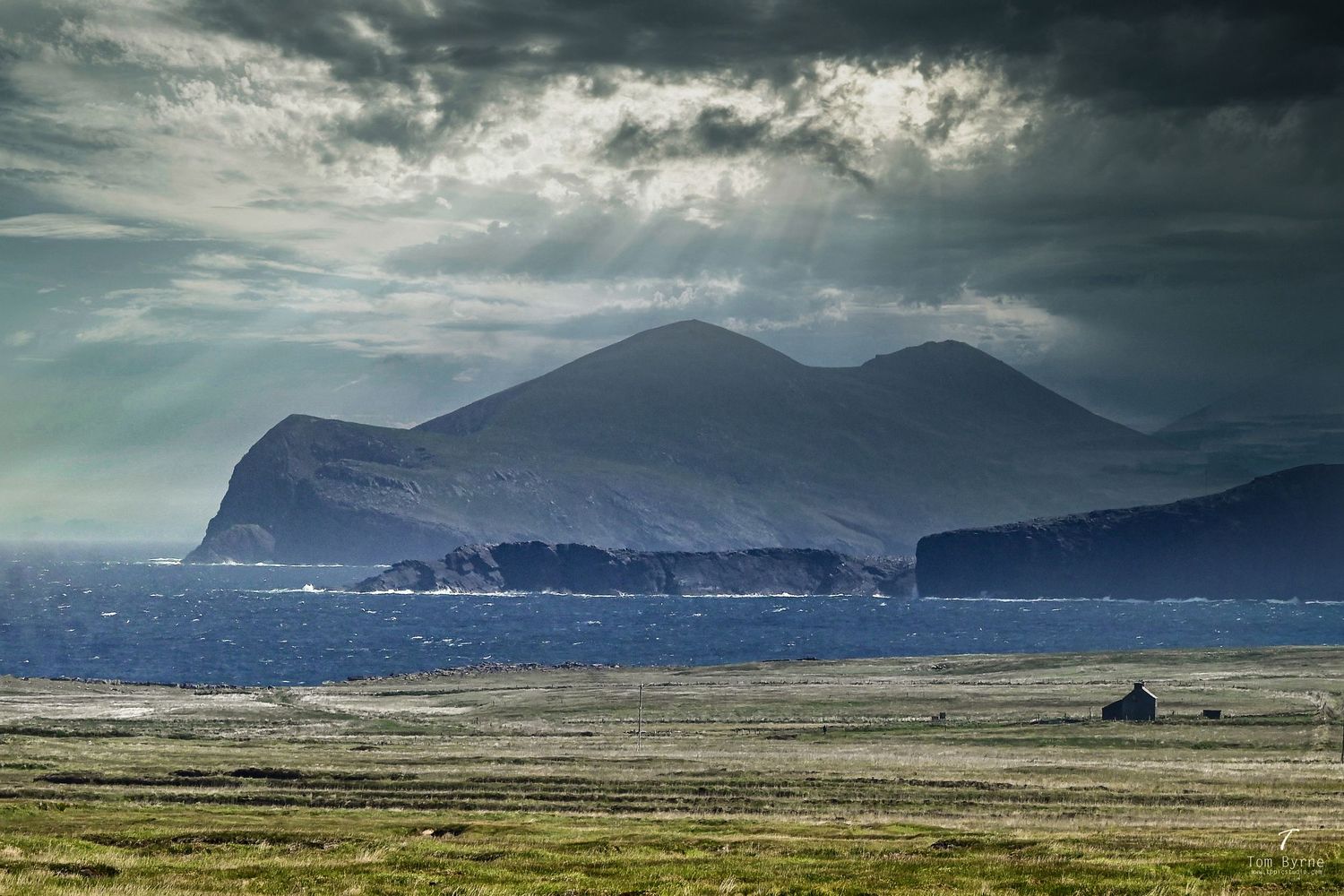 Dramatic coastal landscape with misty mountains, choppy sea, and stormy skies over a rugged green plain.