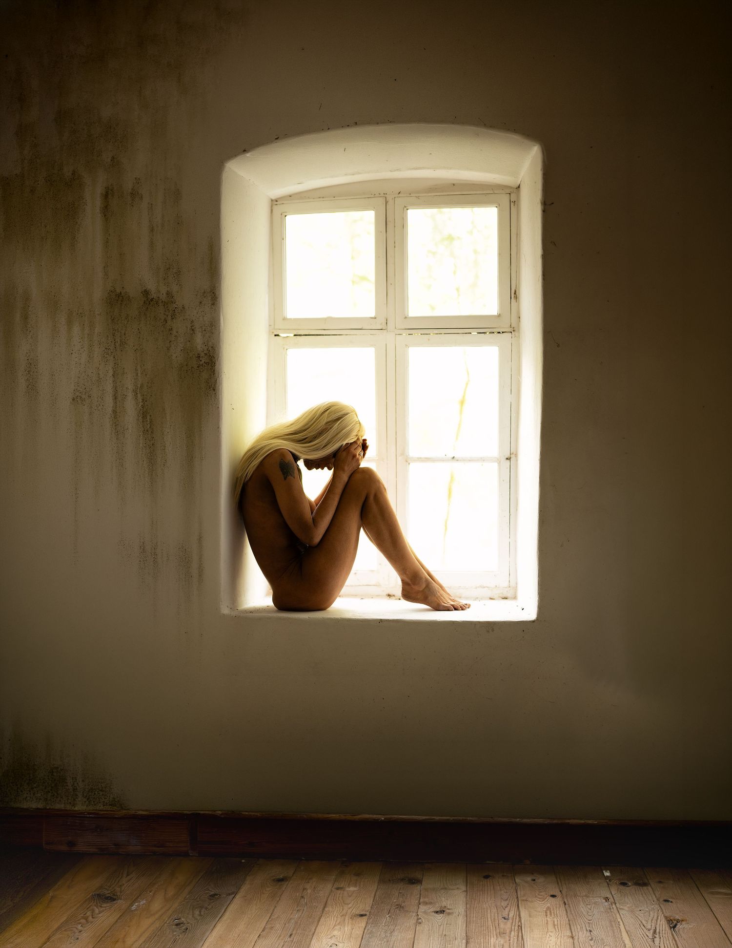 A nude woman sits curled up in a vintage arched window alcove, bathed in soft natural light.