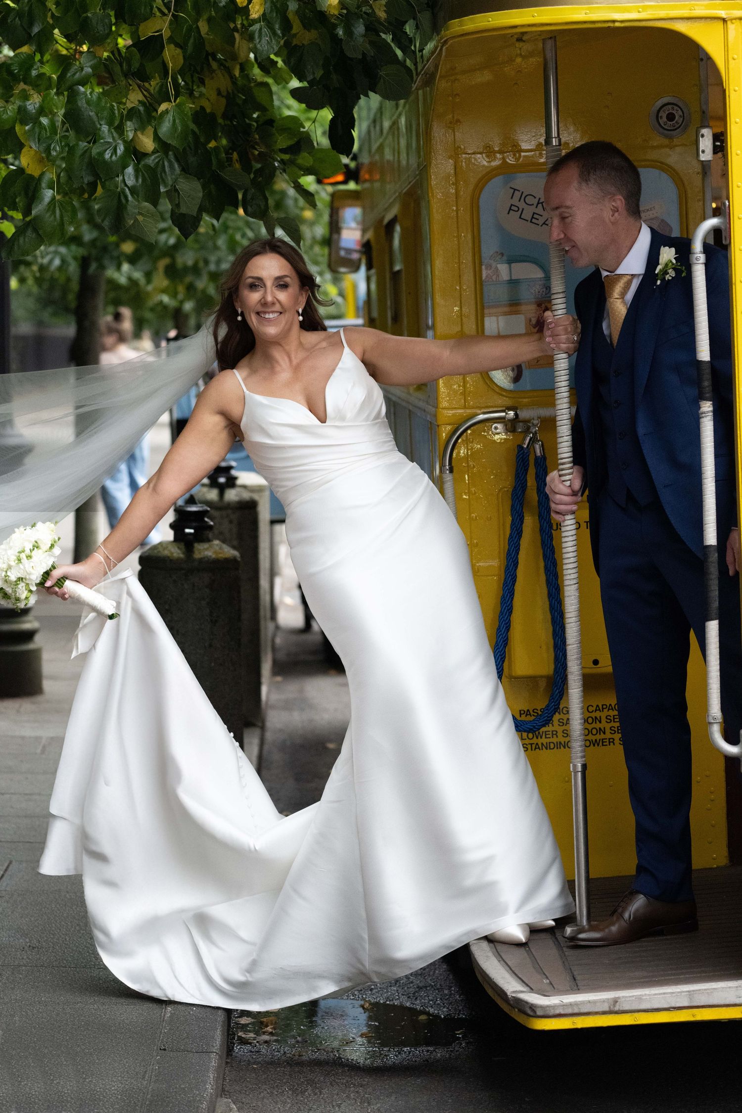 Smiling bride in white gown stepping off yellow tram with groom in navy suit behind her on wedding day.