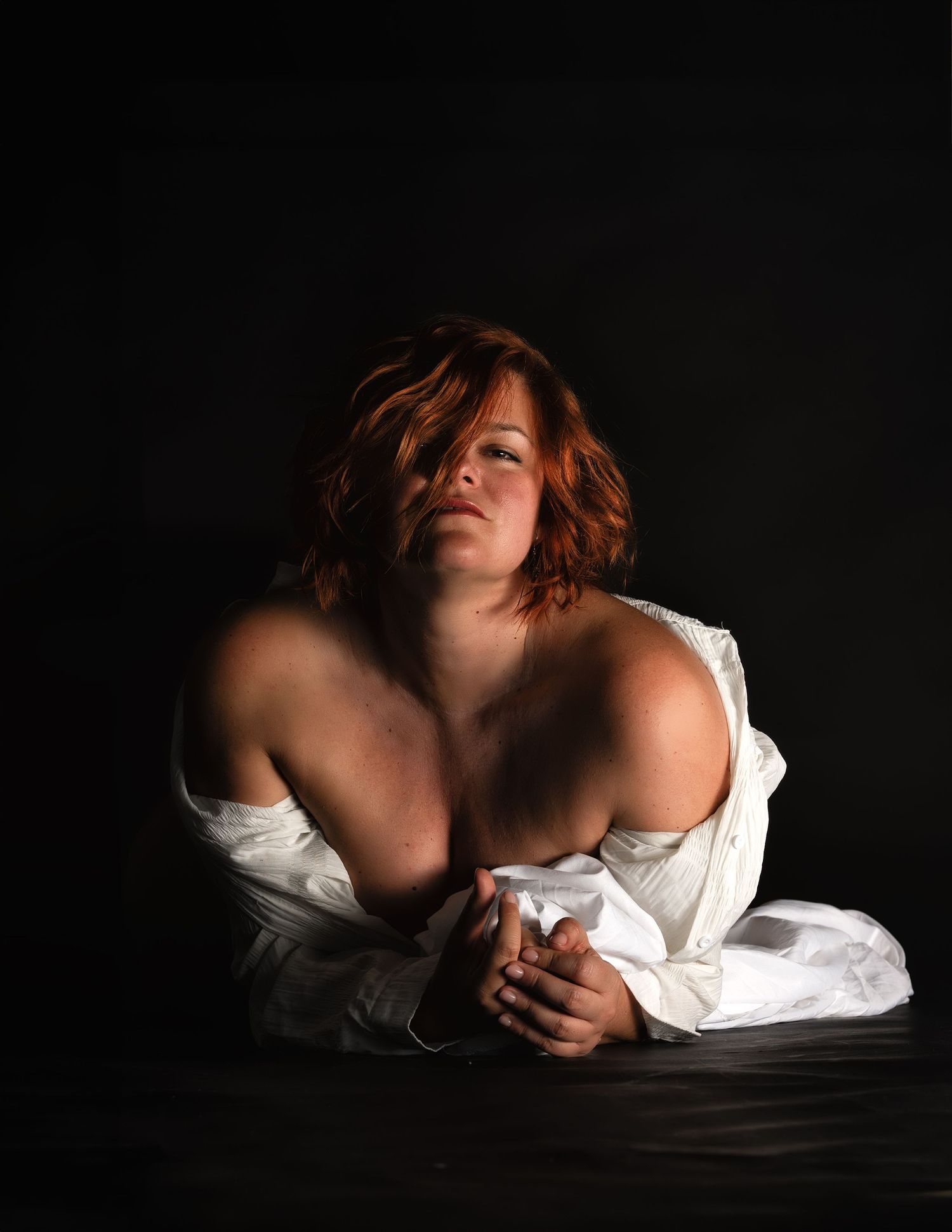 A red-haired woman draped in white fabric poses dramatically against a dark background in artistic portrait photography.