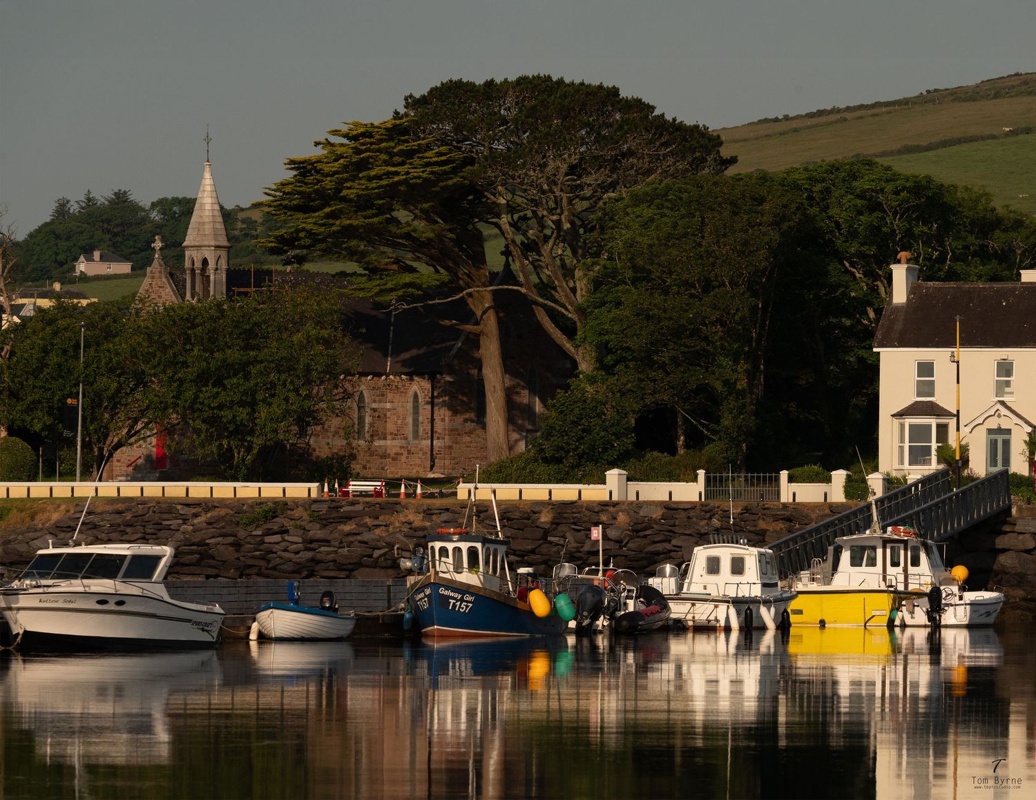 Colorful boats moored in a calm harbor at golden hour, with a church steeple and lush trees in the background.