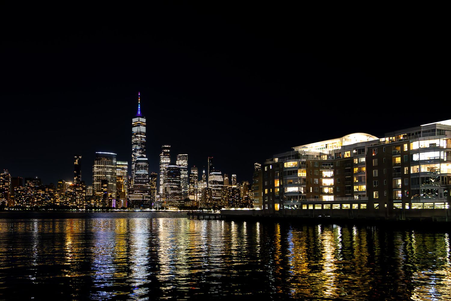New York City skyline at night with One World Trade Center illuminated, viewed across the Hudson River from Jersey City, New Jersey.