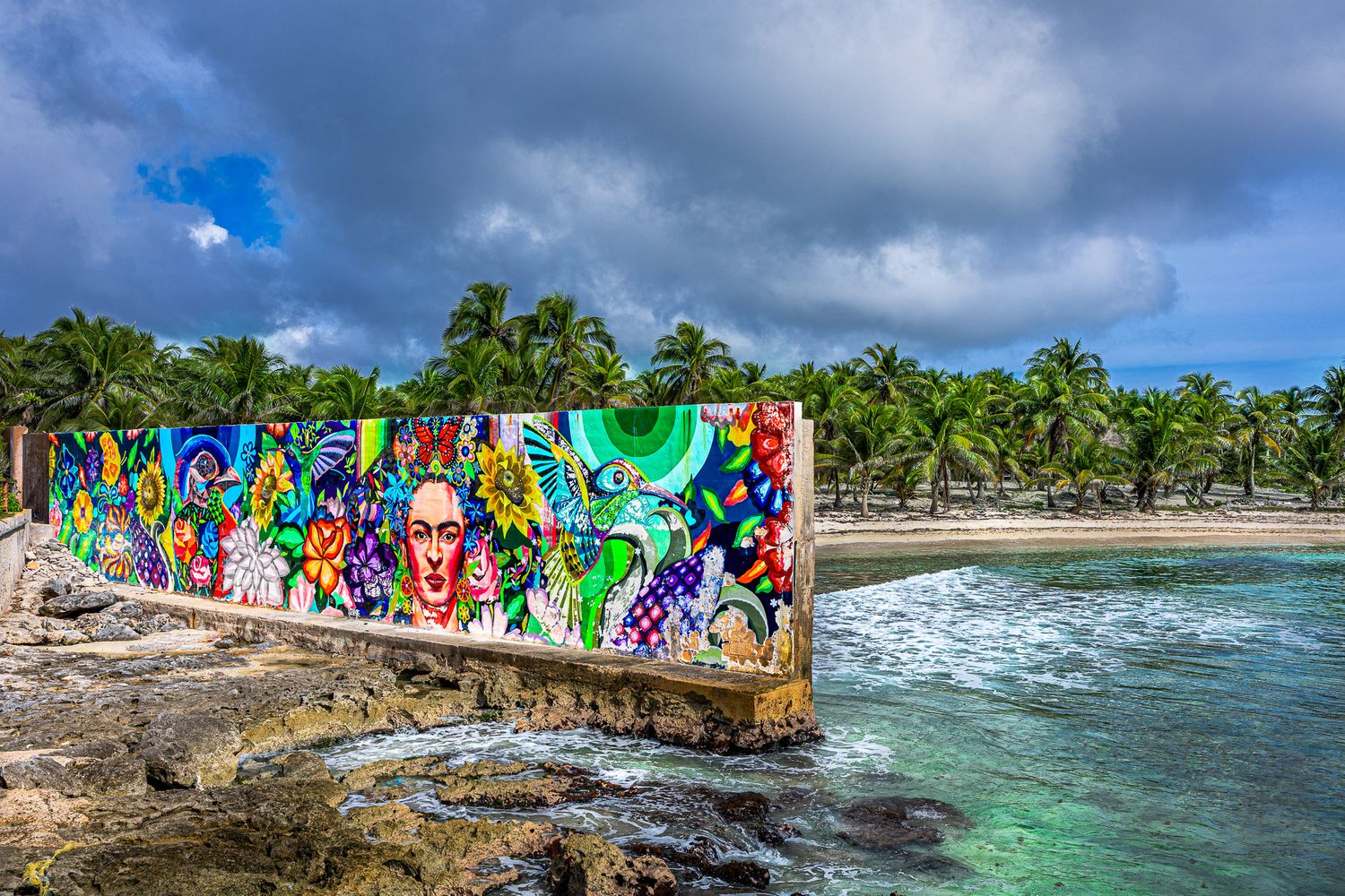 Colorful Frida Kahlo–inspired mural along rocky shoreline at Casa Maya, Mexico with palm trees, turquoise water, and storm clouds.