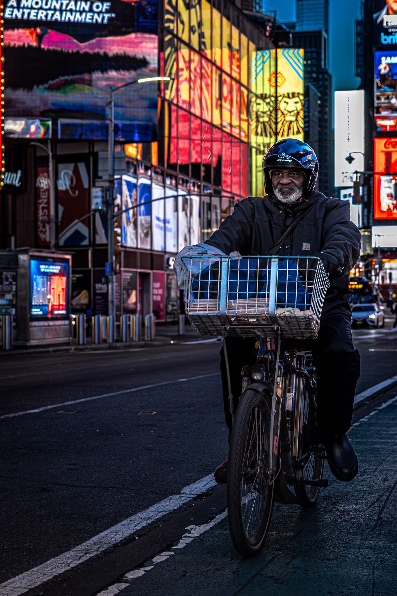 Candid street photograph of a man riding a bicycle through Times Square at night with illuminated billboards in New York City.