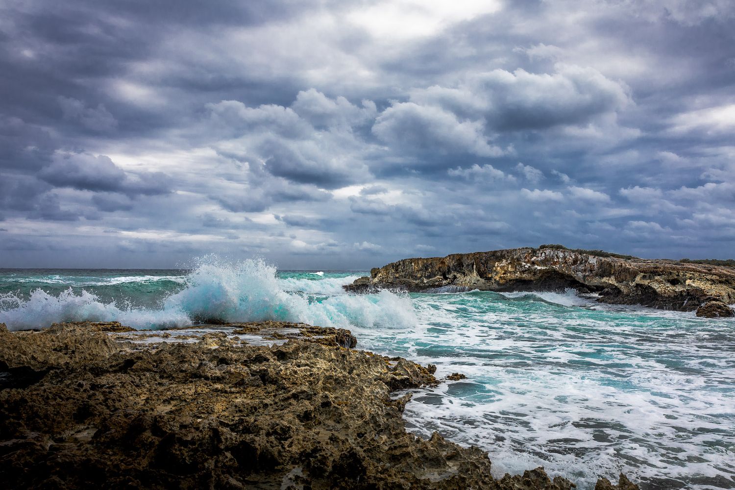 Waves crashing against rocky coastline in Cozumel, Mexico under dramatic storm clouds and turquoise Caribbean water.