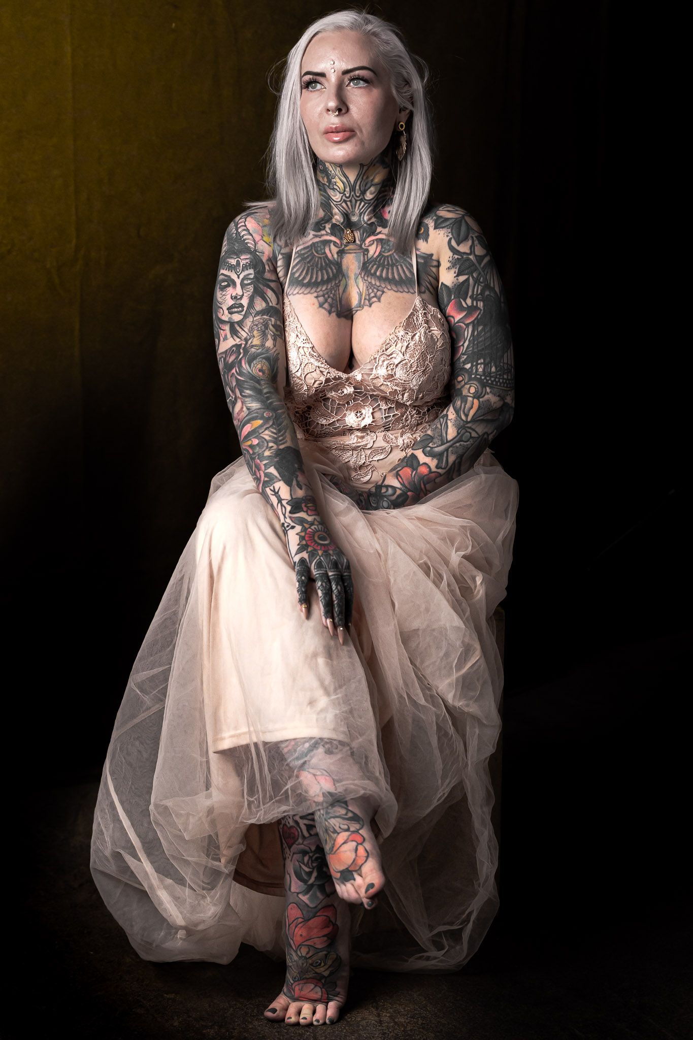 Studio portrait of a tattooed actress in a neutral lace dress seated against a dark background. during a Unique Photo photography event in N