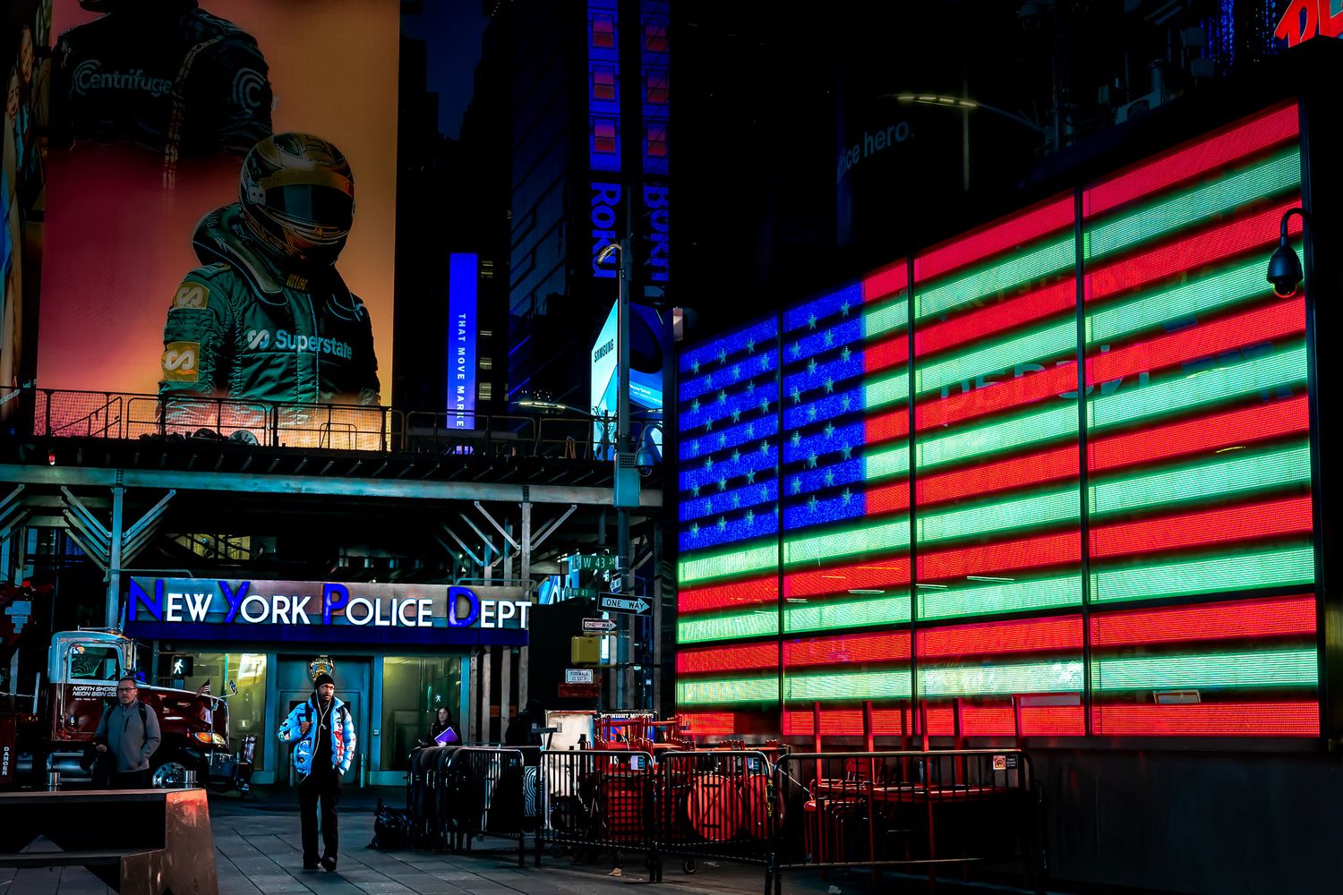 Street photography in Times Square showing the illuminated American flag near NYPD signage at night.