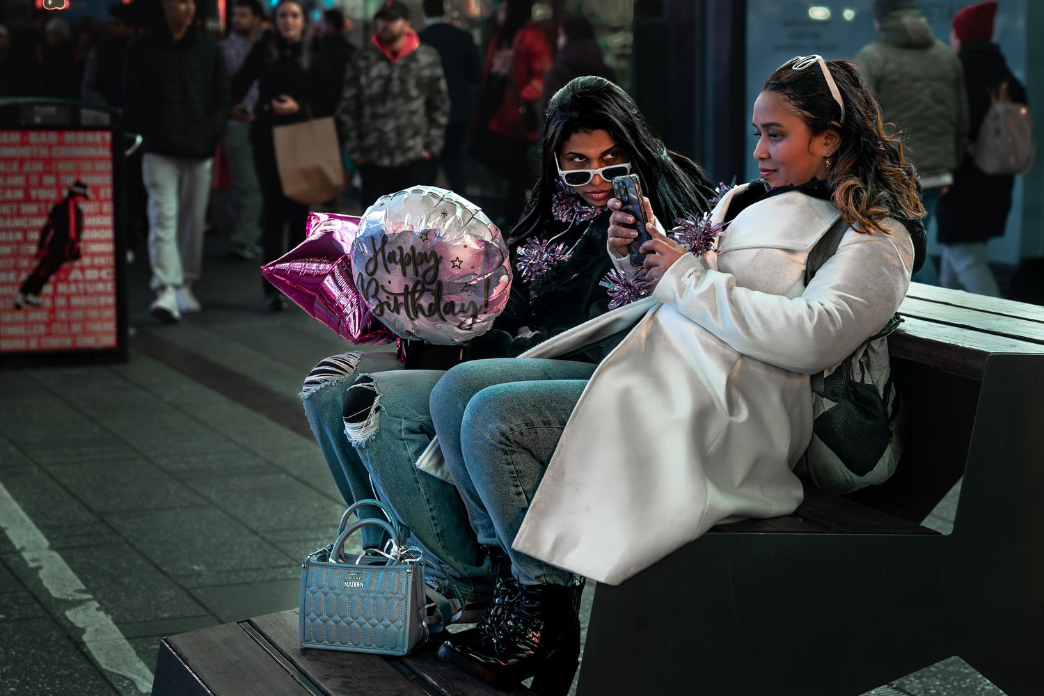Candid street photograph of two women seated with a birthday balloon amid nighttime crowds in Times Square, New York City.