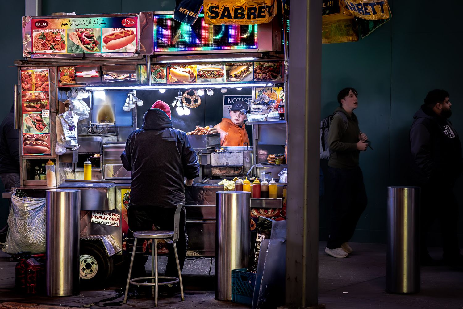 Candid street photograph of a late-night food cart with customers and bright signage in Times Square, New York City.