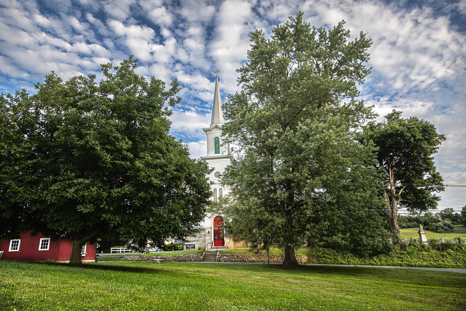 Historic white church in Hope, New Jersey framed by large trees and green lawn under a dramatic cloud-filled summer sky.
