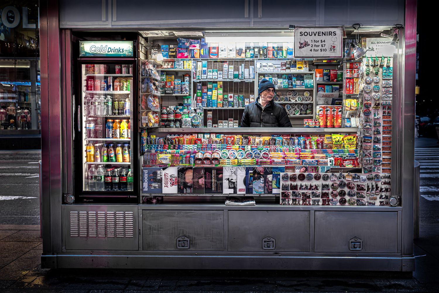 Candid street photograph of a newsstand vendor inside a brightly lit kiosk at night in Times Square, New York City.