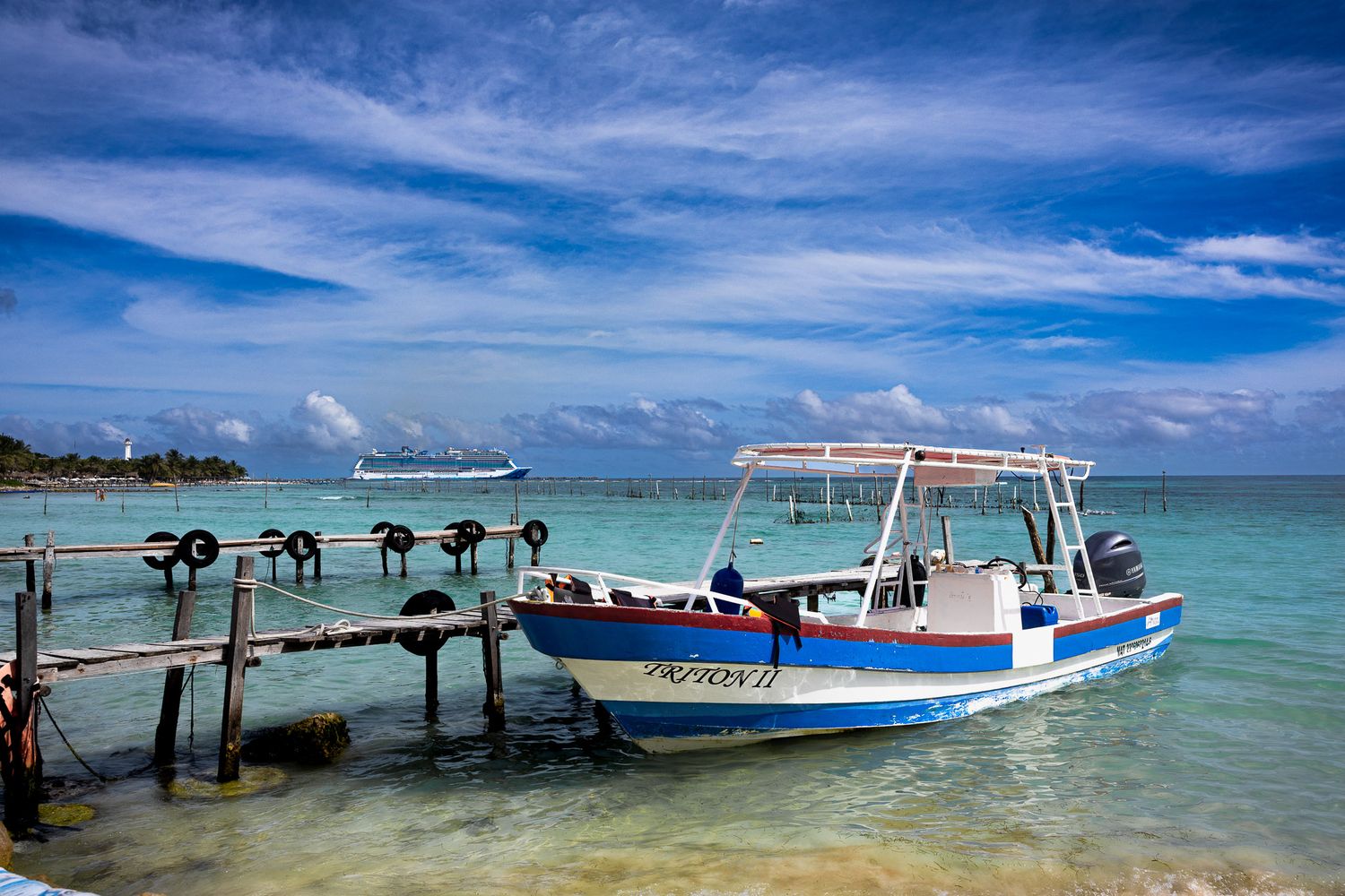 Small fishing boat docked in turquoise water at Casa Maya, Mexico with wooden pier and Norwegian Bliss cruise ship offshore.
