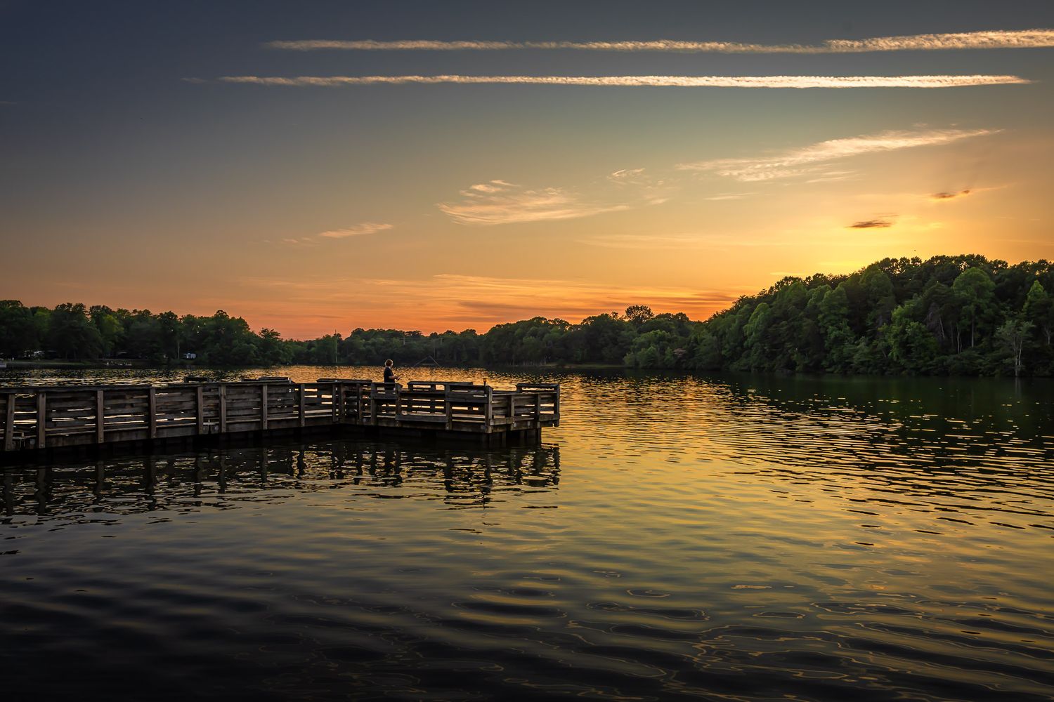 Sunset over a calm lake in High Point, North Carolina, with a lone person fishing from a wooden pier and golden reflections on the water.