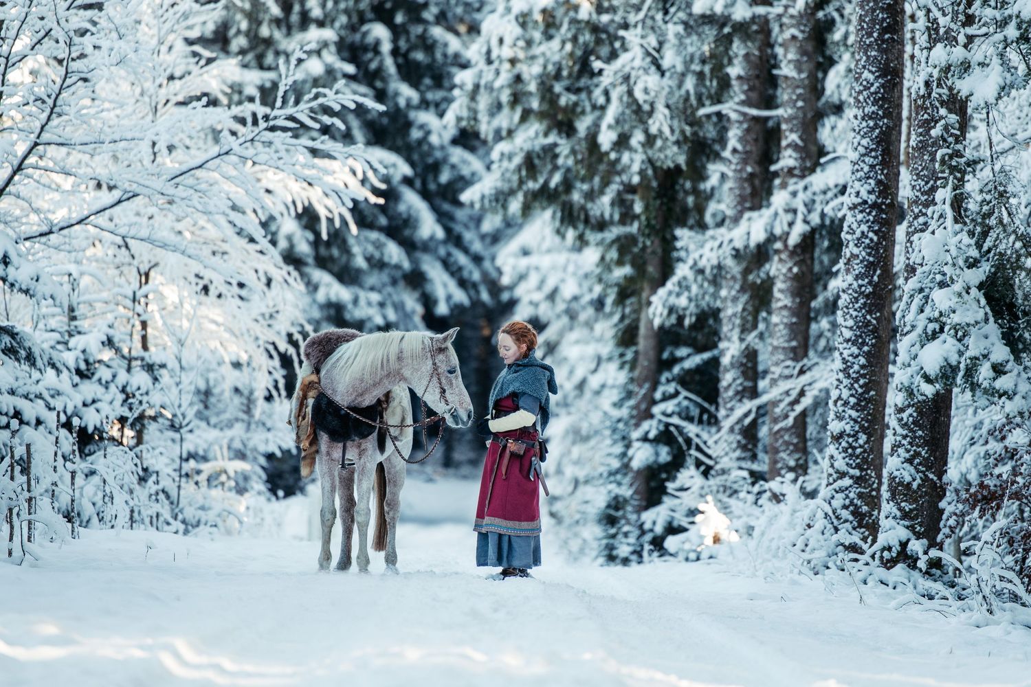Grey horse and person in traditional dress walking through snow-covered forest.