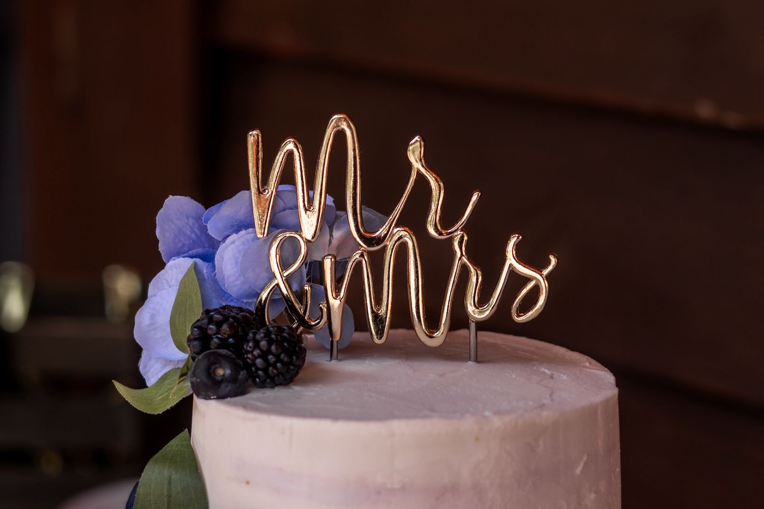 Elegant wedding cake with gold wire Mr. & Mrs. topper, purple hydrangea and blackberry decorations on white frosting.
