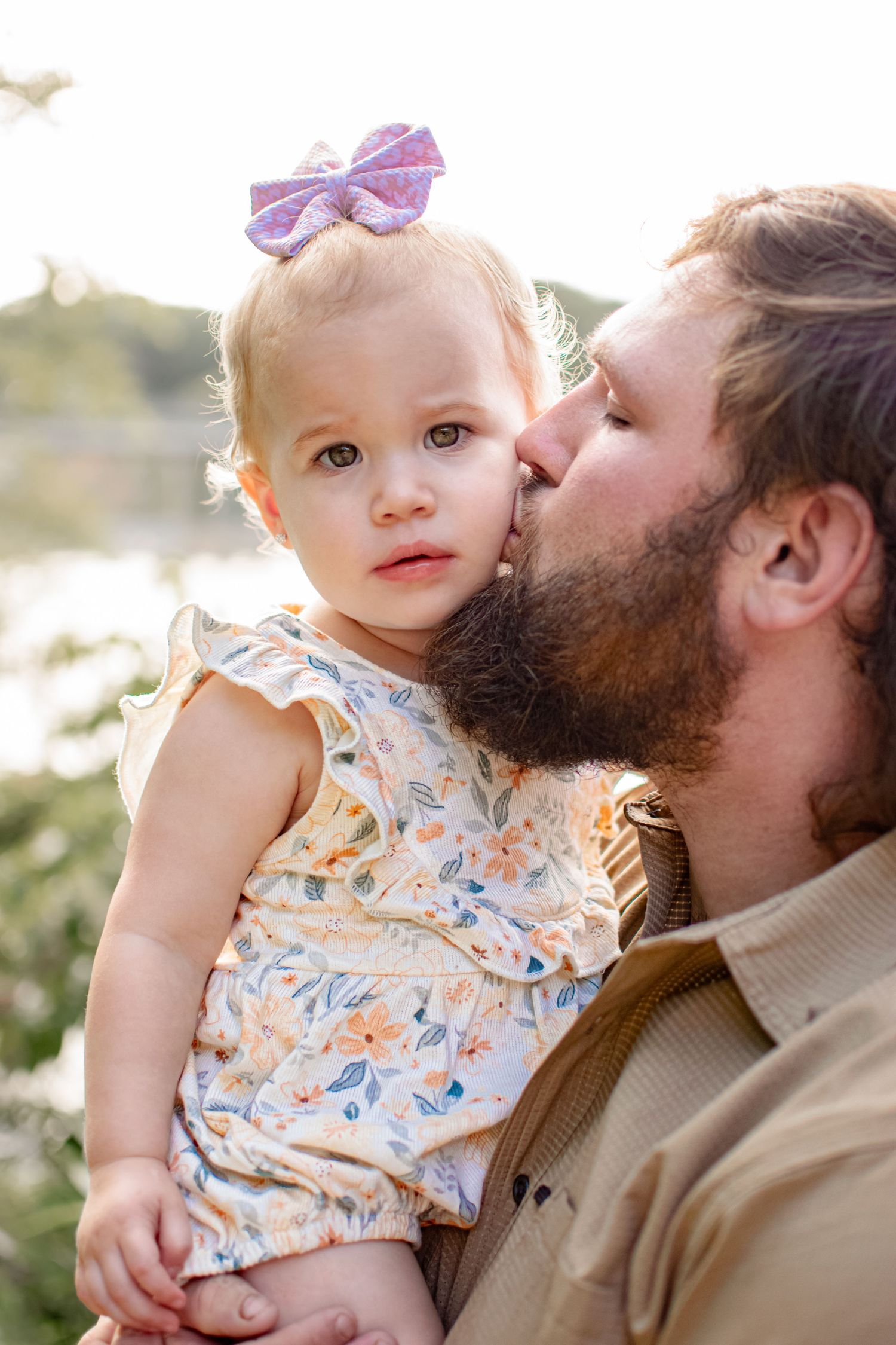 A bearded father gently kisses the cheek of his baby girl wearing a floral outfit and purple bow outdoors.