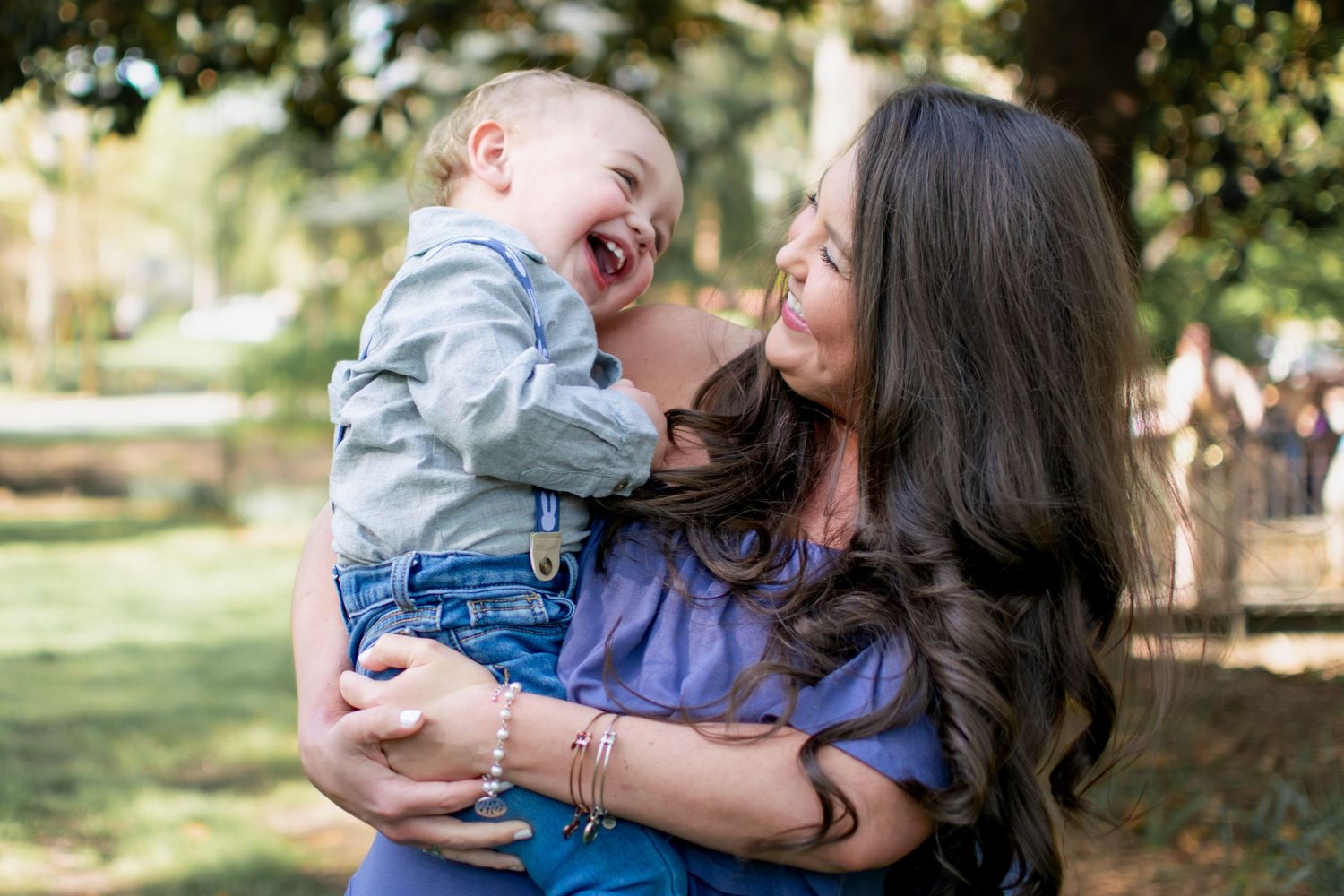 Joyful dark-haired mother holding laughing baby boy outdoors in green park setting.