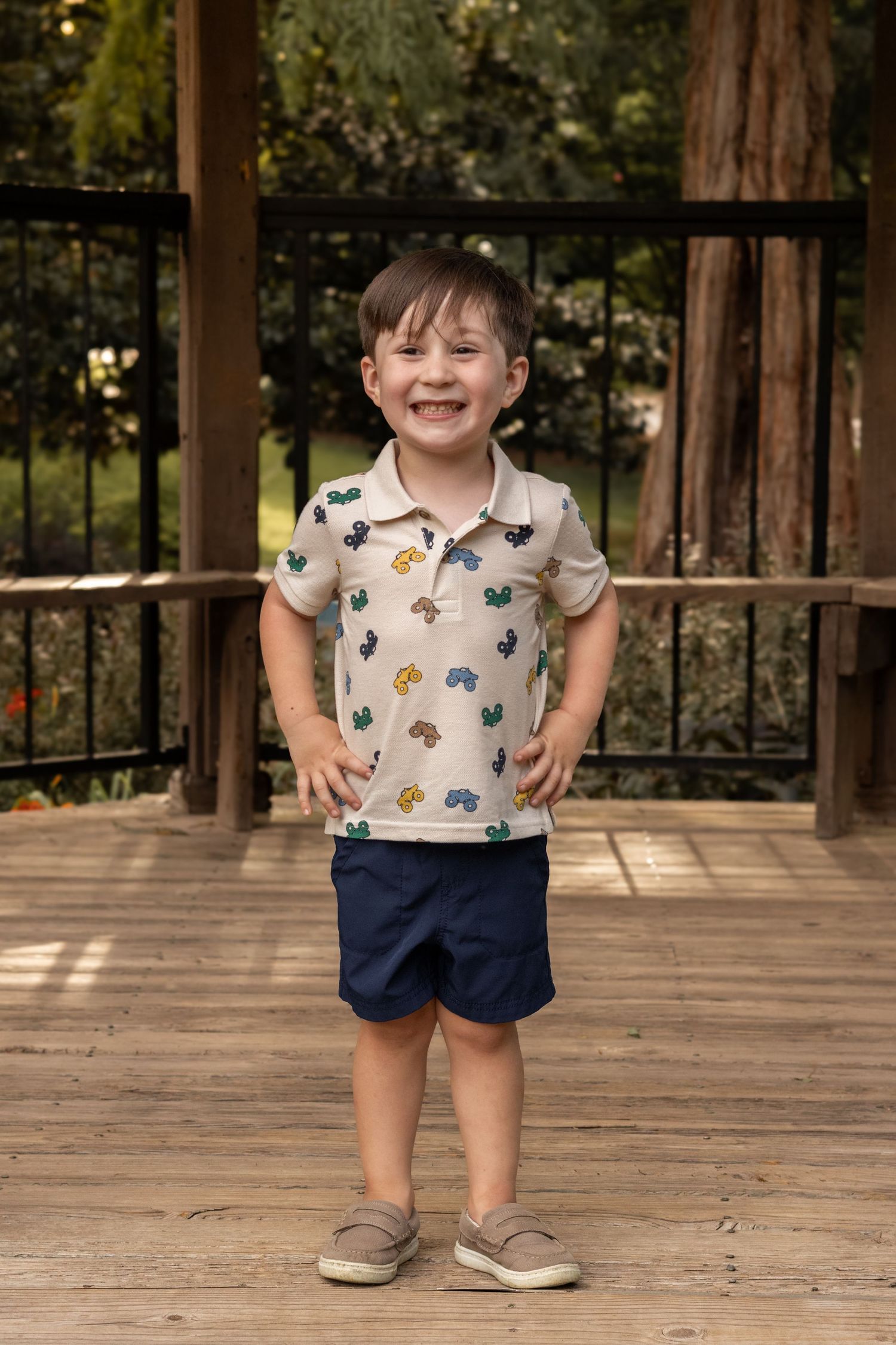 Smiling young boy in a patterned polo shirt and navy shorts standing on a wooden deck outdoors.