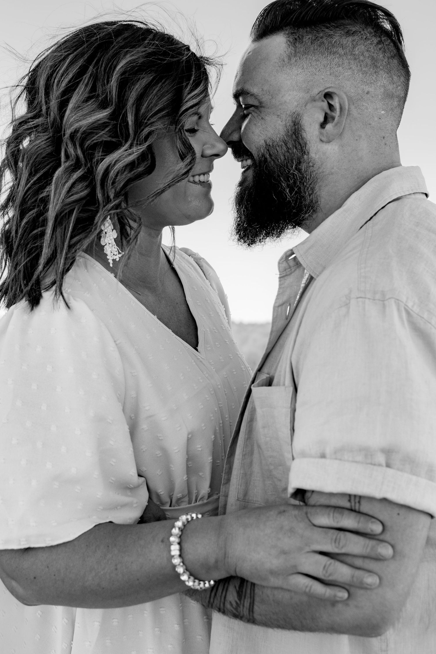 Black and white photo of a smiling couple embracing closely, the woman in a white dress and man with a beard.