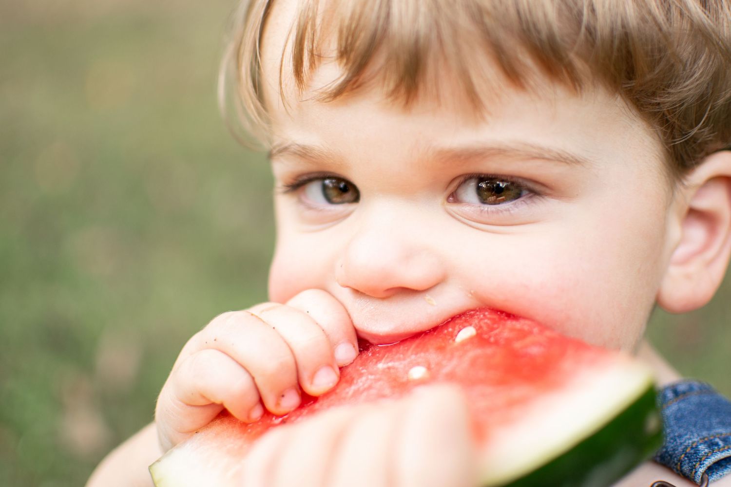 A cute toddler with big brown eyes takes a bite of a fresh watermelon slice outdoors.
