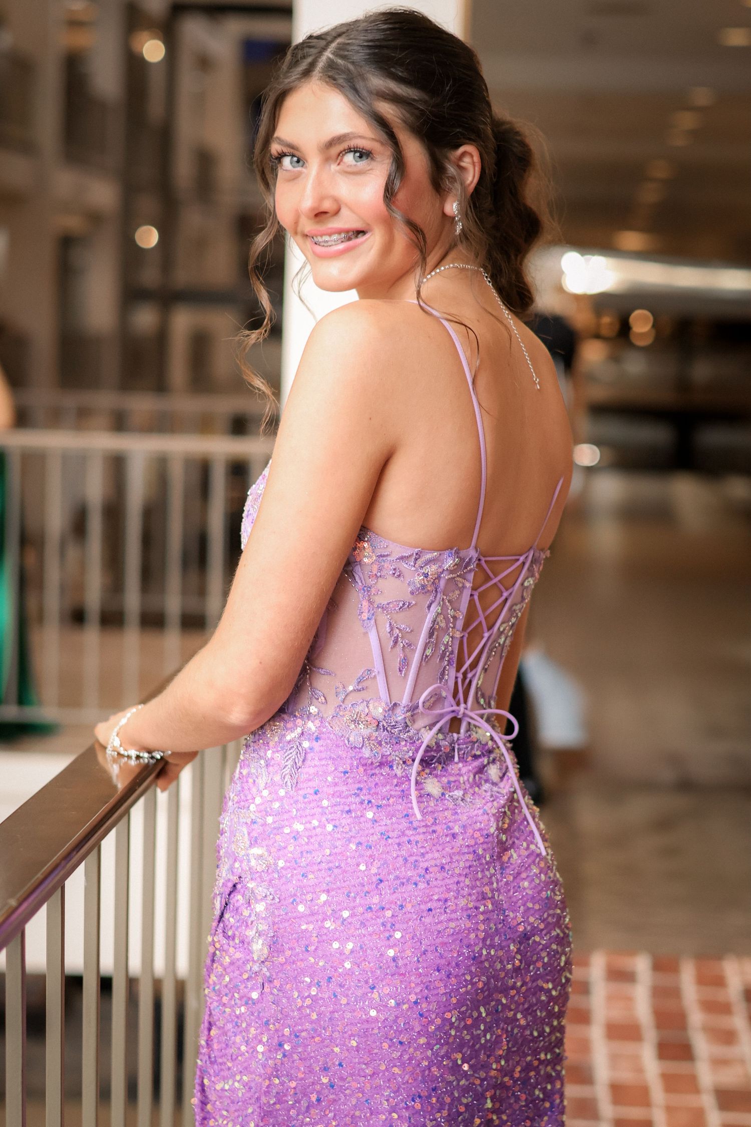 A young woman in a sparkly lavender backless prom dress with lace-up corset detail smiles over her shoulder.