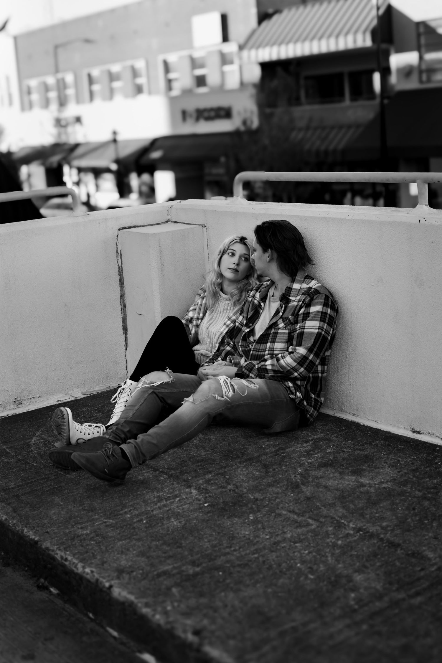 Black and white photo of a romantic couple sitting together against a wall on an urban rooftop, wearing plaid shirts.