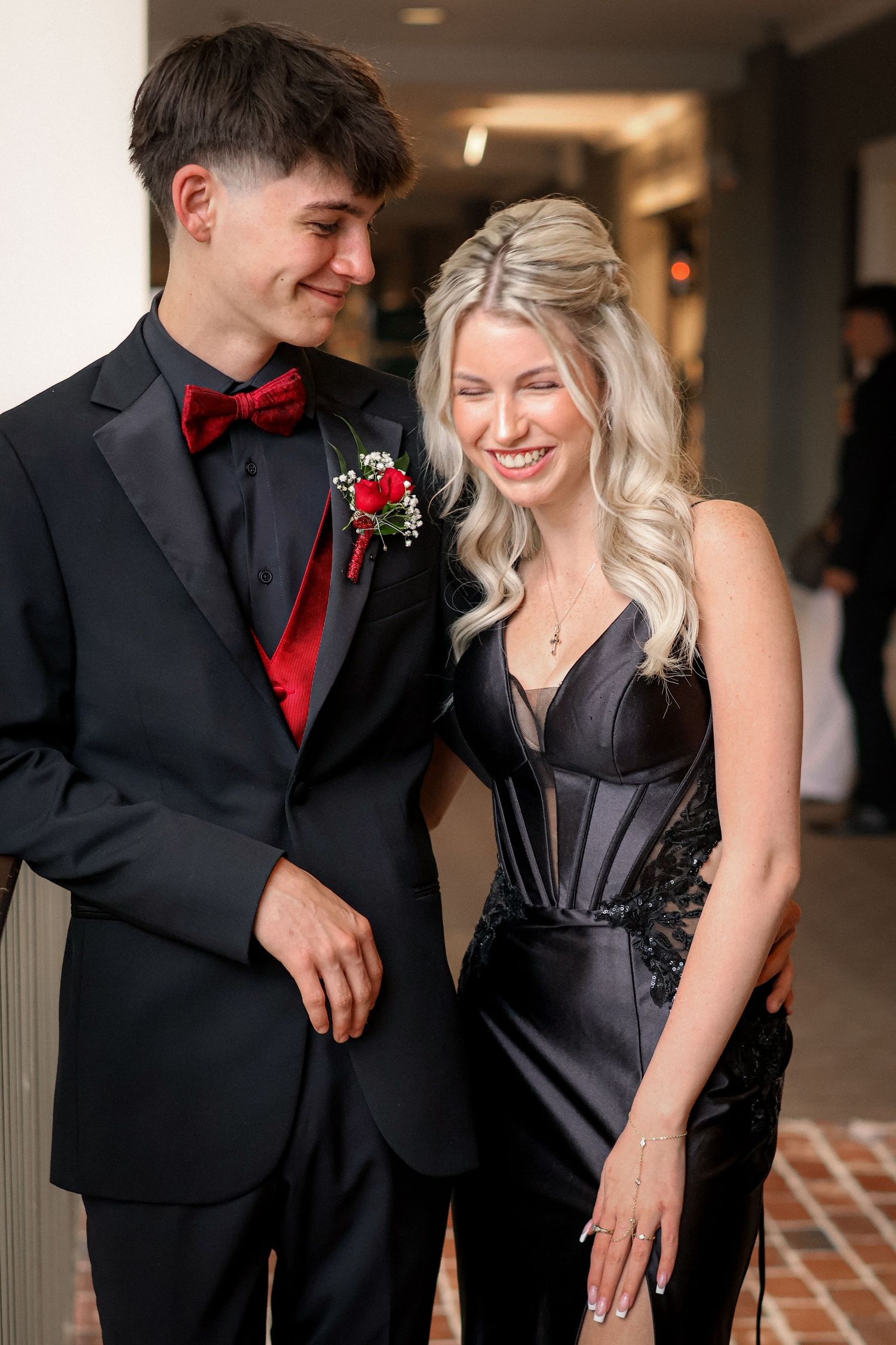 Elegant prom couple in matching black formal wear with red boutonniere laughing together indoors.