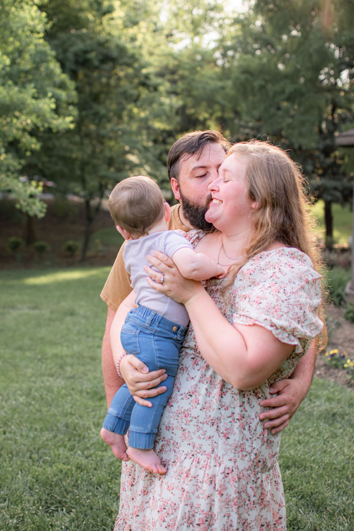 A bearded man and woman share a kiss while holding their baby together in a lush green backyard.