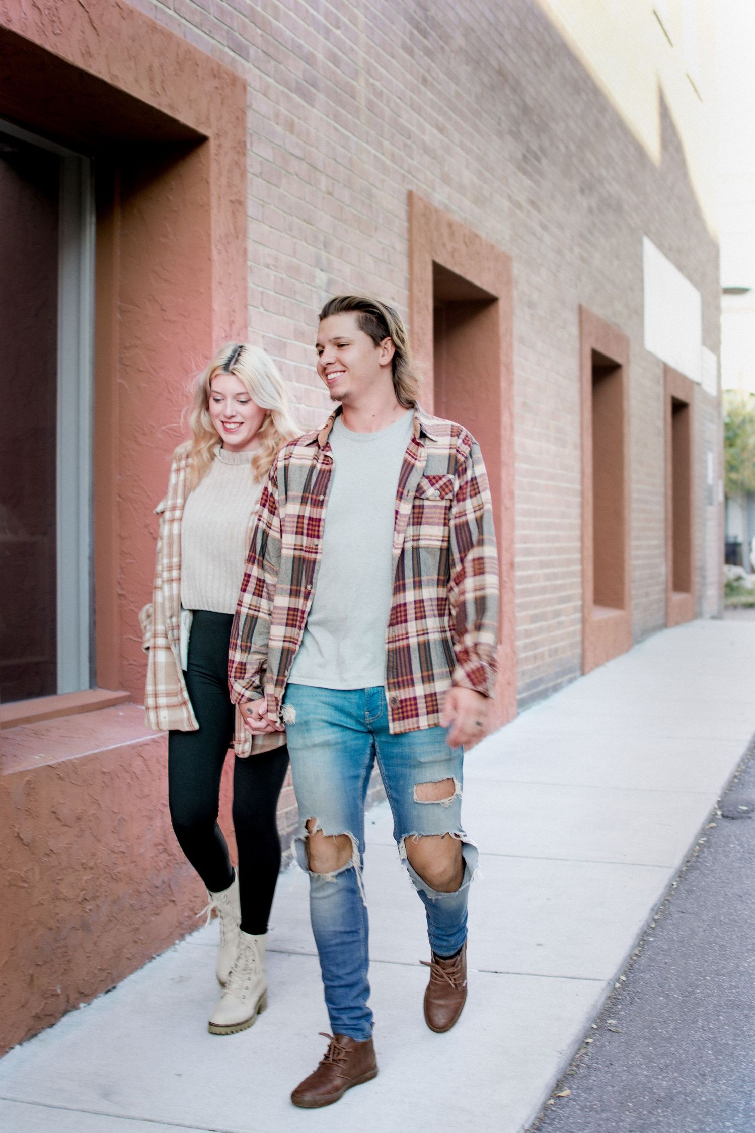 Smiling couple in matching plaid flannel shirts holding hands while walking along brick building sidewalk.