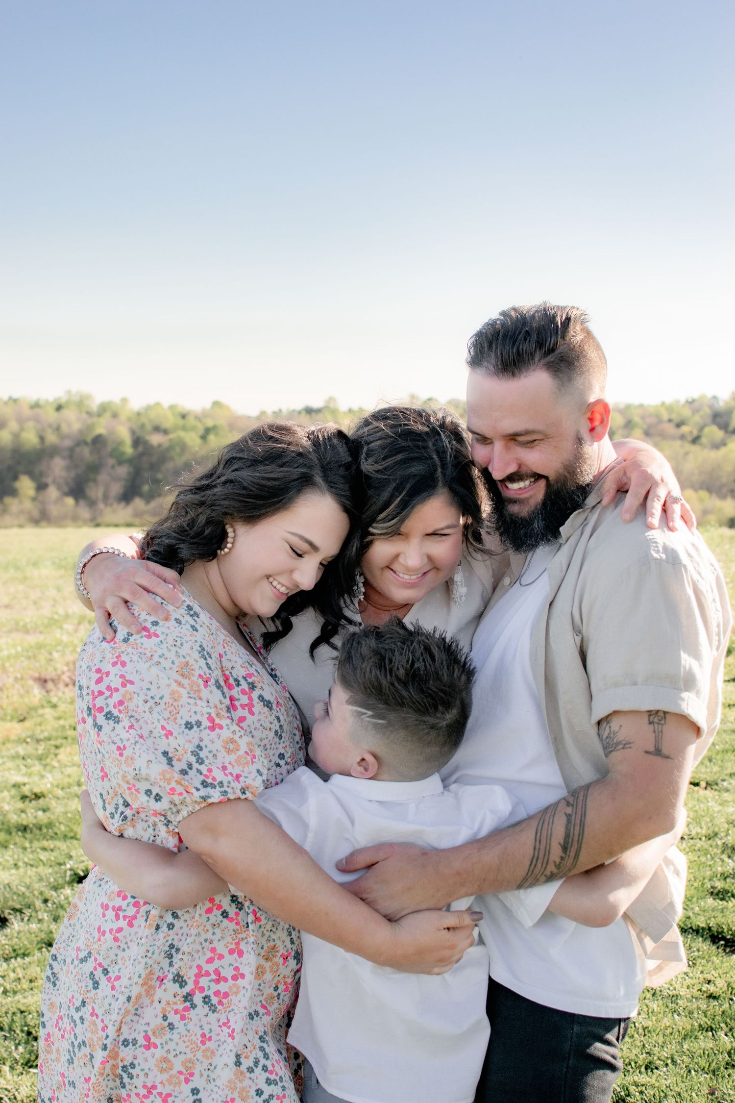 Joyful family of four sharing a warm group hug in an open green field under a clear blue sky.