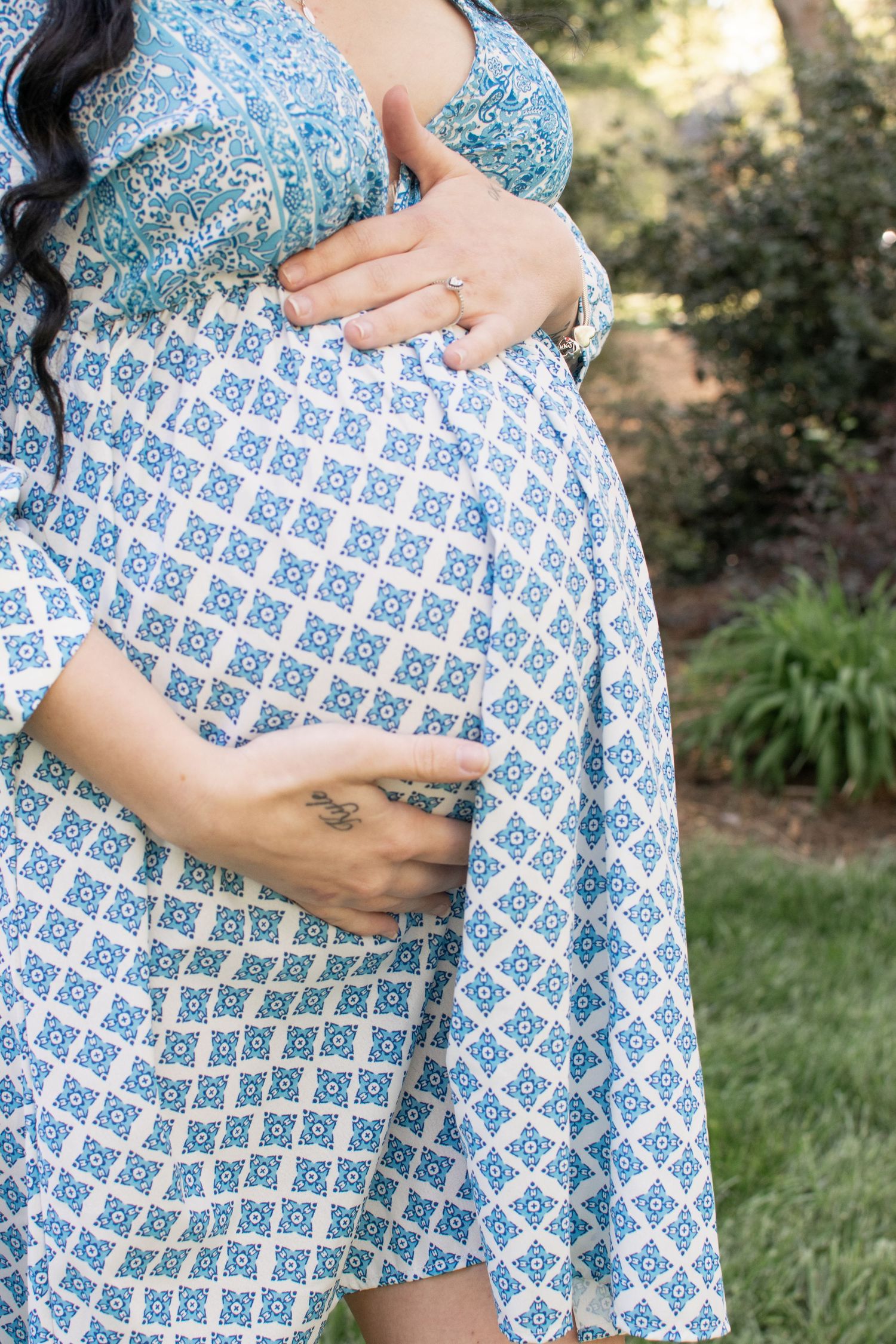 Close-up of pregnant woman in blue patterned dress cradling baby bump with both hands outdoors.