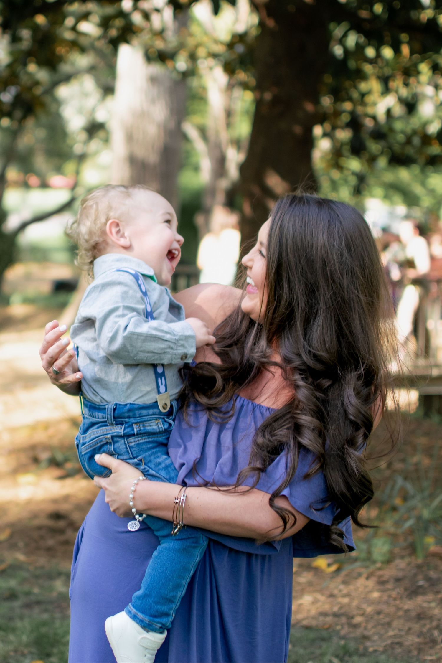 Joyful mother in purple dress holding laughing baby boy under shaded trees in park.