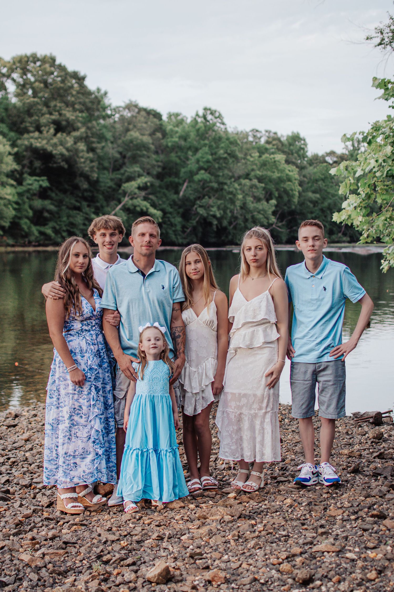 A large family of seven poses together on a rocky riverbank surrounded by lush green trees.