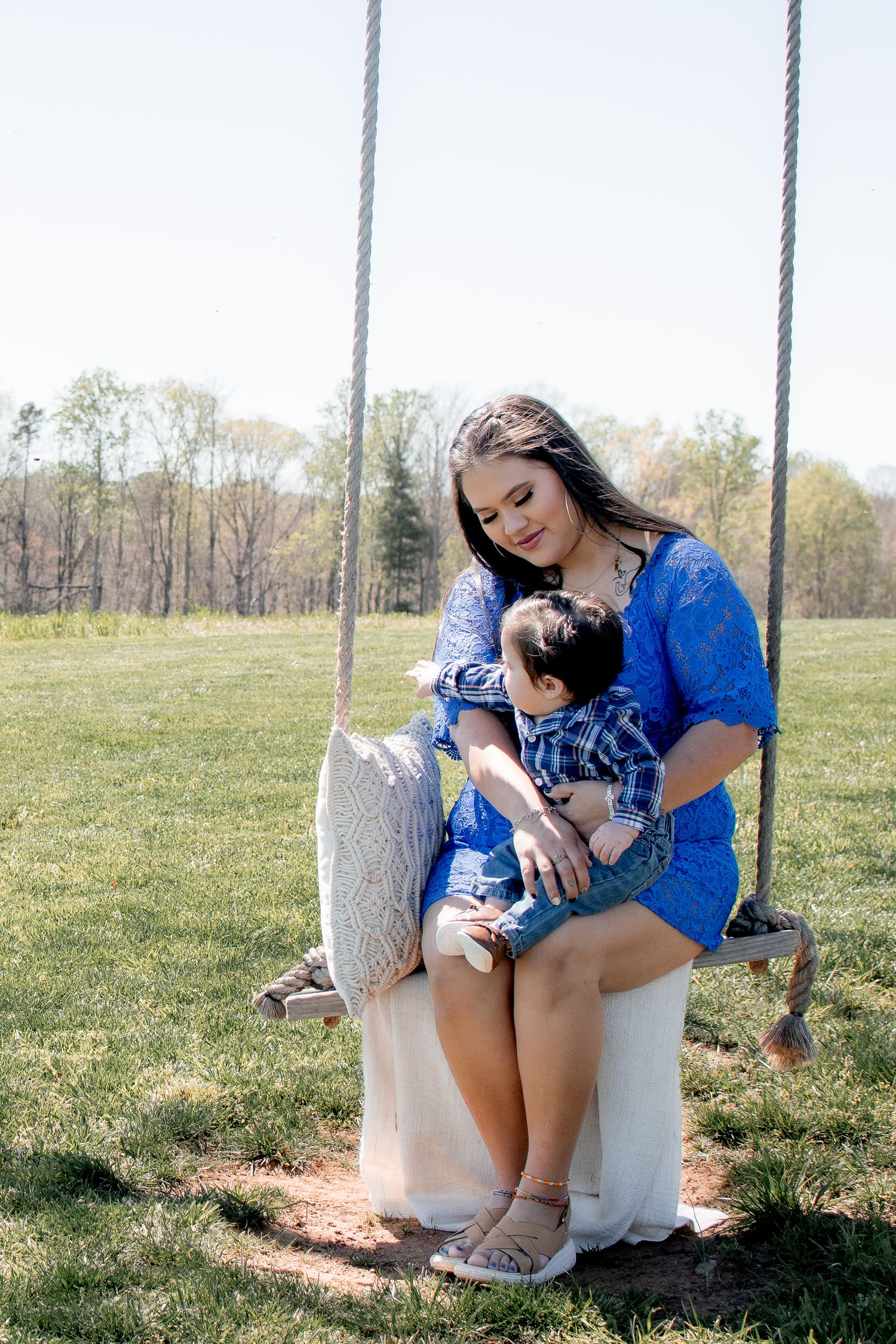 Mother in blue lace dress sitting on rope swing holding young child in sunny meadow.