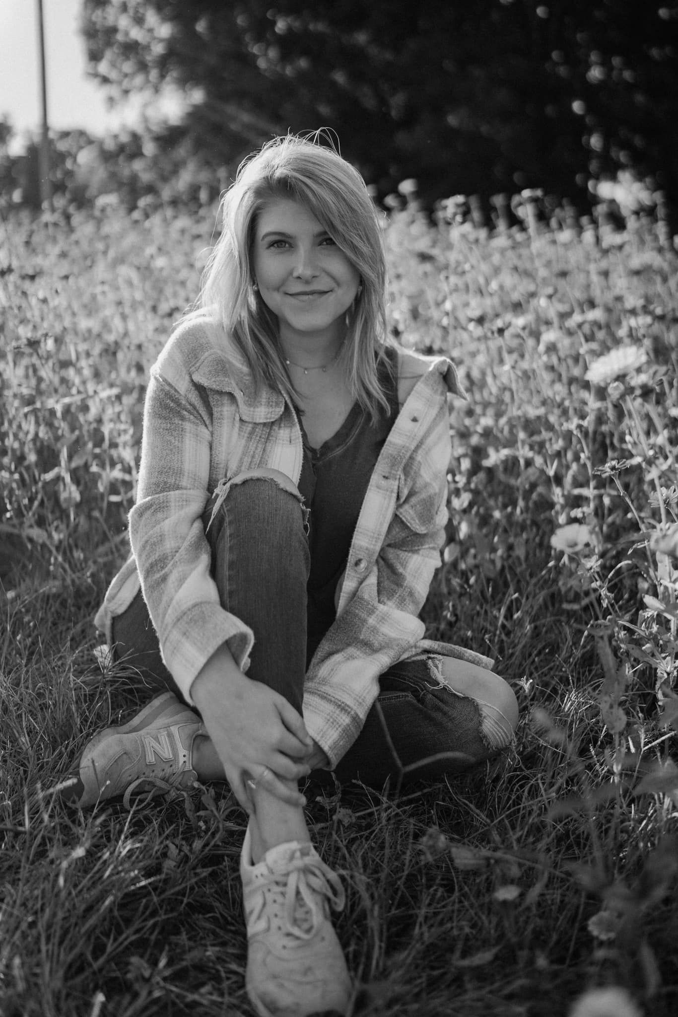 Black and white photo of young blonde woman sitting in wildflower field wearing plaid shirt.