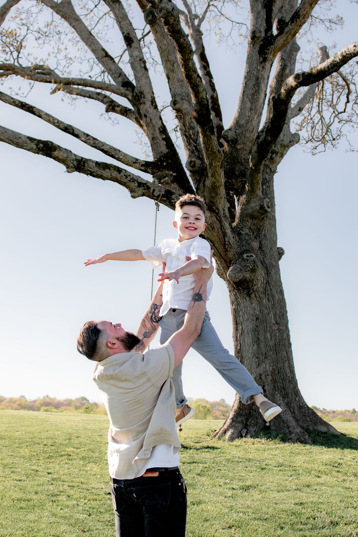 A joyful father lifts his laughing son into the air outdoors beneath a large bare tree on a sunny day.